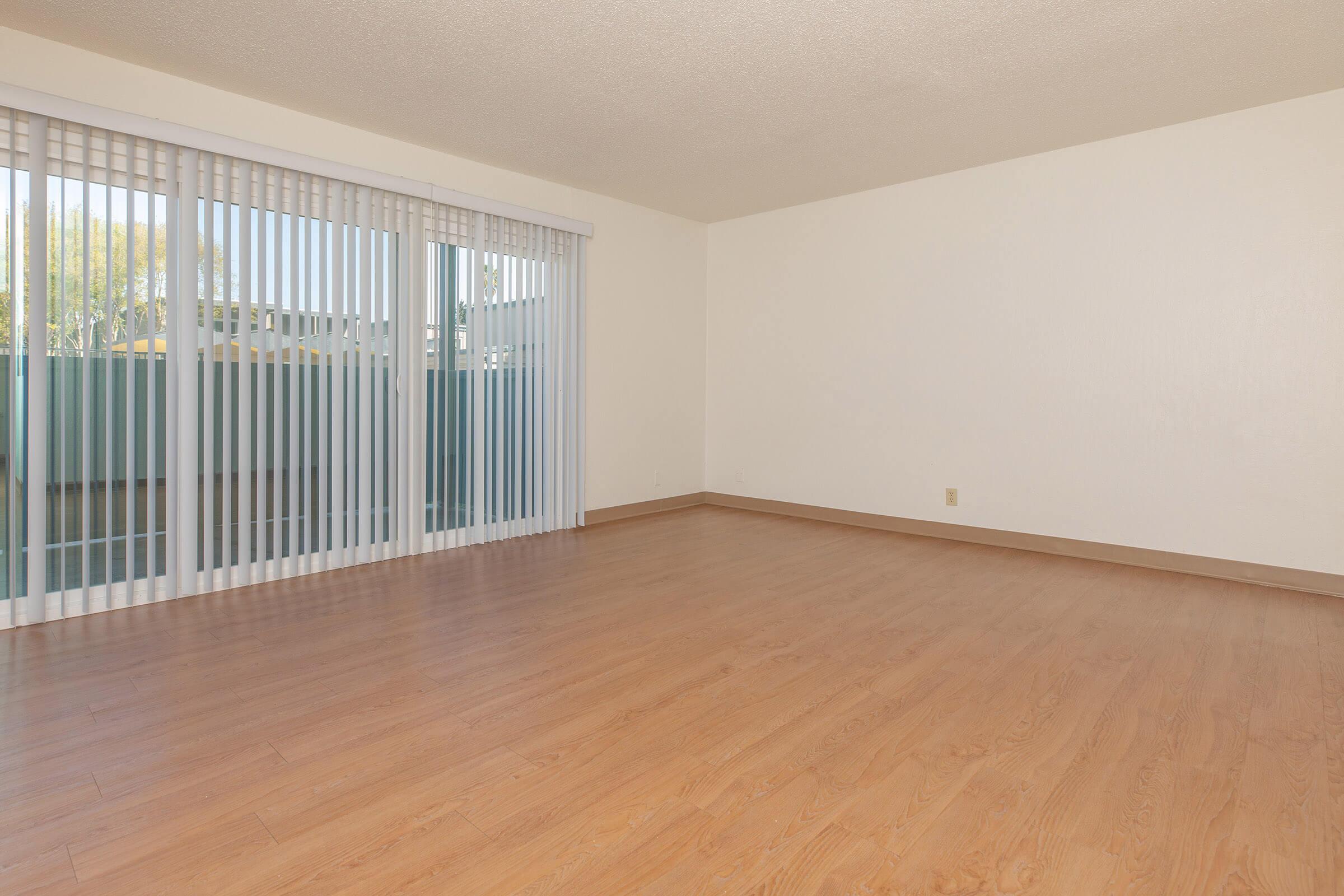 Empty room with light-colored walls and wooden flooring. Large sliding glass doors with vertical blinds open to an outdoor area. The space is well-lit with natural light, creating a bright and clean atmosphere. No furniture or decorations present.