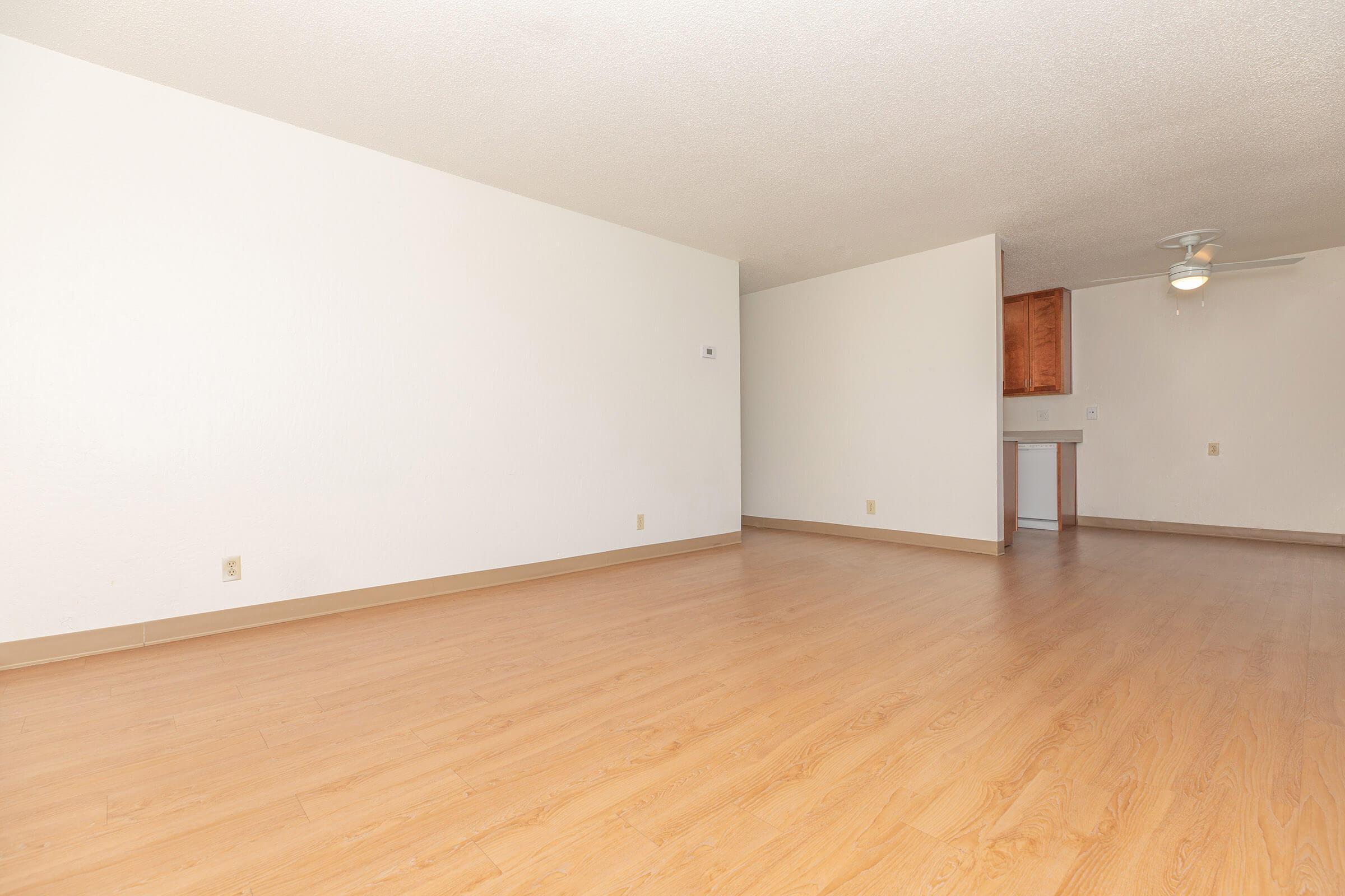 Spacious, empty room with light wood flooring and white walls. A ceiling fan is visible in the corner, and a small kitchenette area is partially seen in the background. The room has a bright, open feel, suitable for various furniture arrangements.