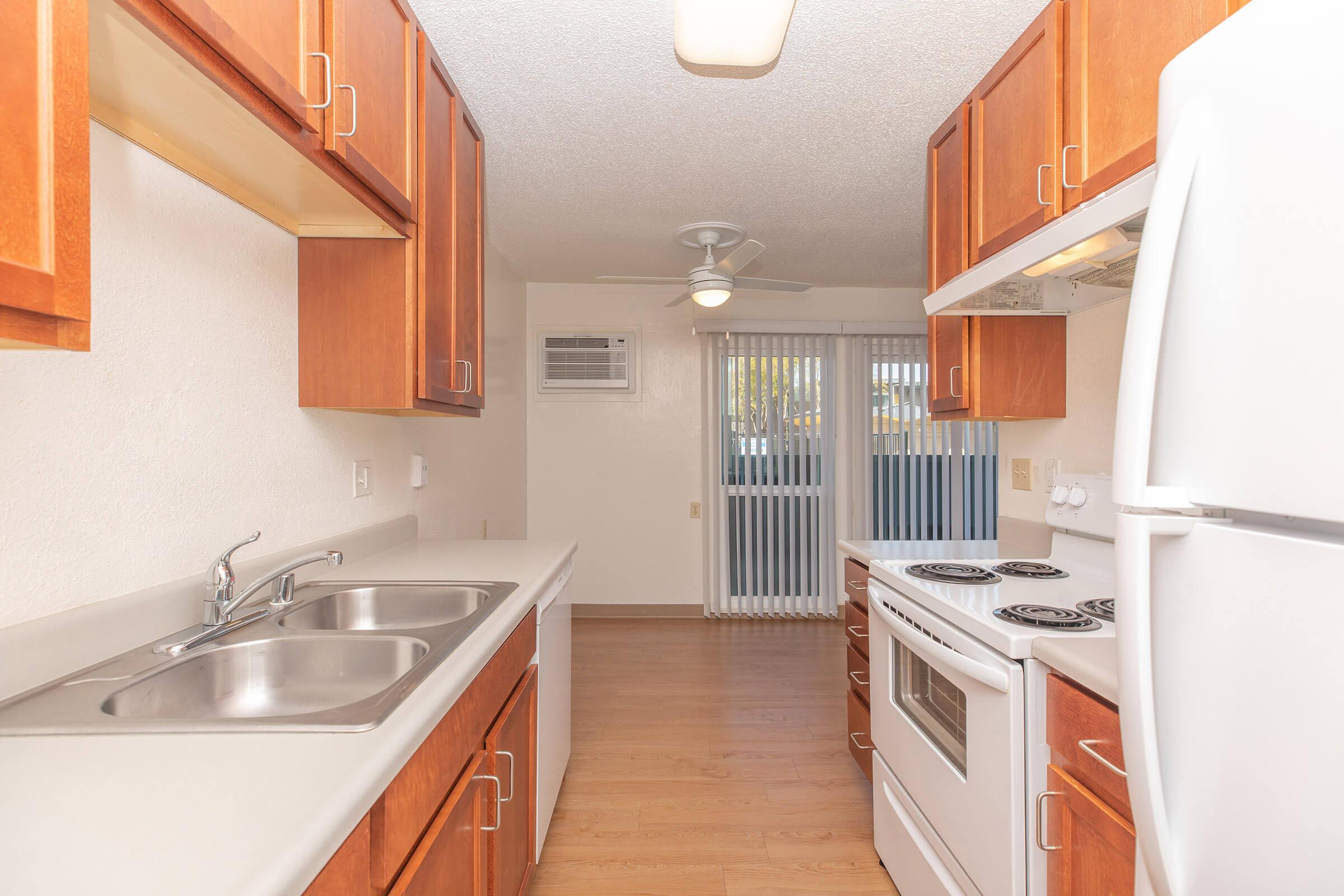A modern kitchen featuring wooden cabinetry, a double sink, a refrigerator, an oven with a stovetop, and a ceiling fan. There are large windows with blinds that allow natural light into the space, and the flooring is light wood laminate. An air conditioning unit is visible on the wall.