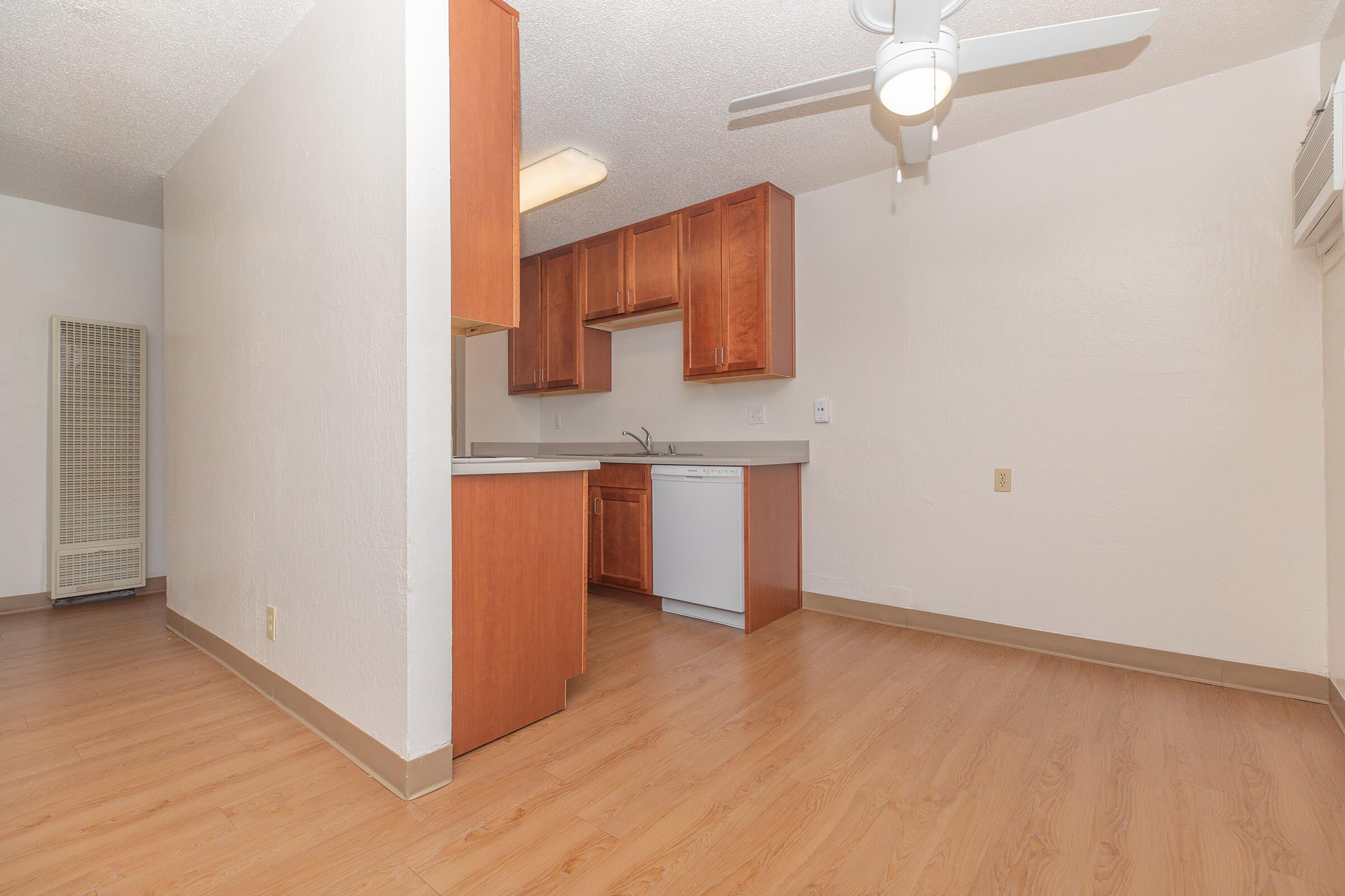 Interior view of a small kitchen area featuring wooden cabinets, a countertop, a dishwasher, and a ceiling fan. The flooring is light-colored, and the walls are painted white. A wall-mounted air conditioner is also visible in the space, which has a clean and simple design.