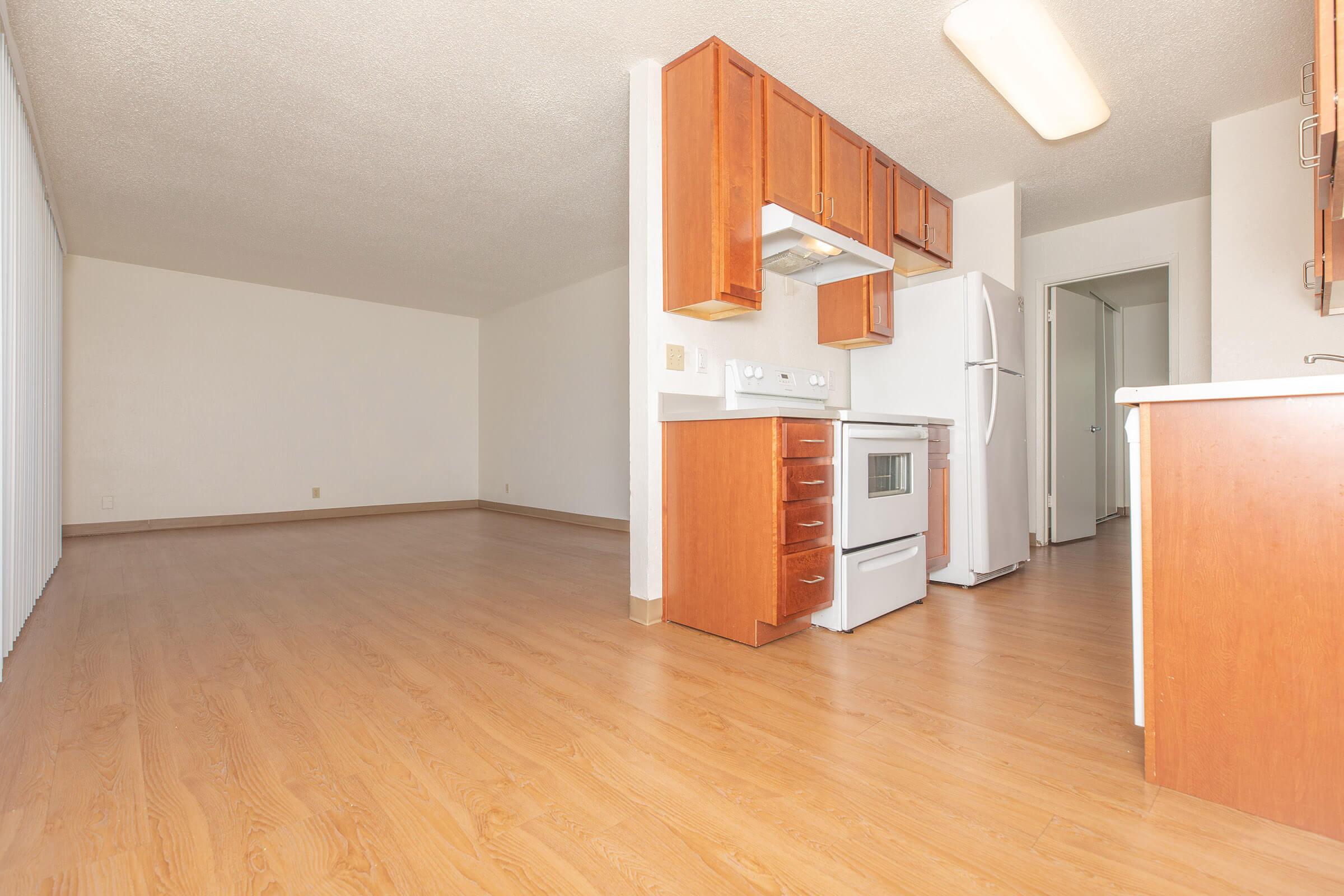 A modern kitchen and living space featuring light wood flooring. The kitchen includes wooden cabinetry, a white refrigerator, a stove, and an oven. Natural light enters from a large window, with neutral-colored walls creating an open and spacious ambiance. The room is empty, suggesting it's ready for new occupants.