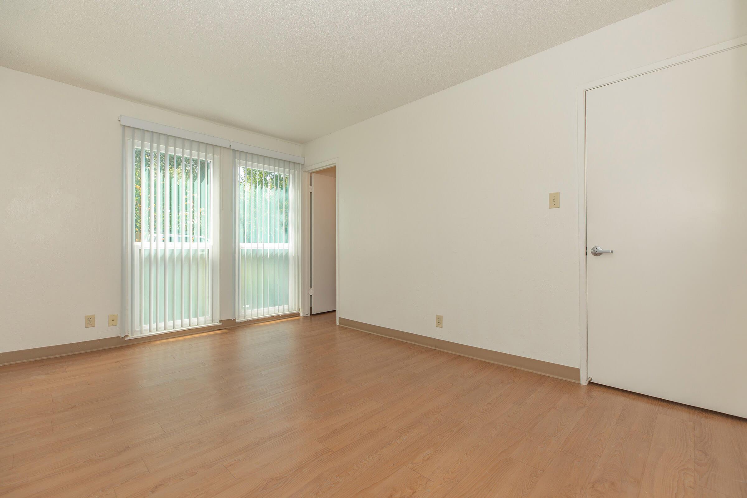 A vacant room with light-colored walls and laminate flooring. Large windows with vertical blinds allow natural light to enter. A door leads to another space, while the overall layout appears spacious and unadorned, providing an opportunity for personalization.
