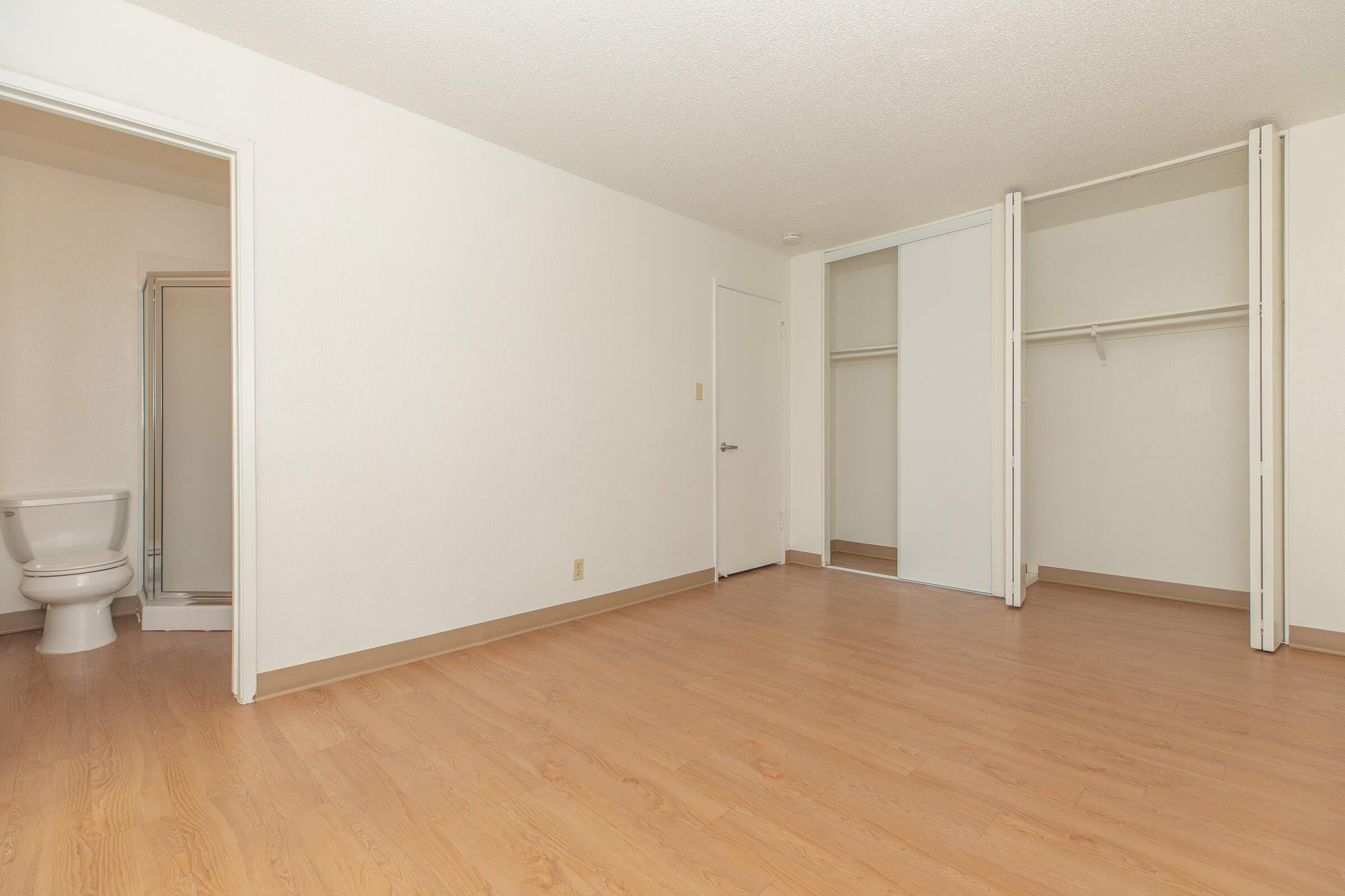 A spacious, empty room with light-colored walls and wooden flooring. On the left side, there’s a small bathroom visible with a toilet and shower area. In the right corner, there are two large sliding closet doors. The overall atmosphere is bright and minimalist, ideal for personalizing.