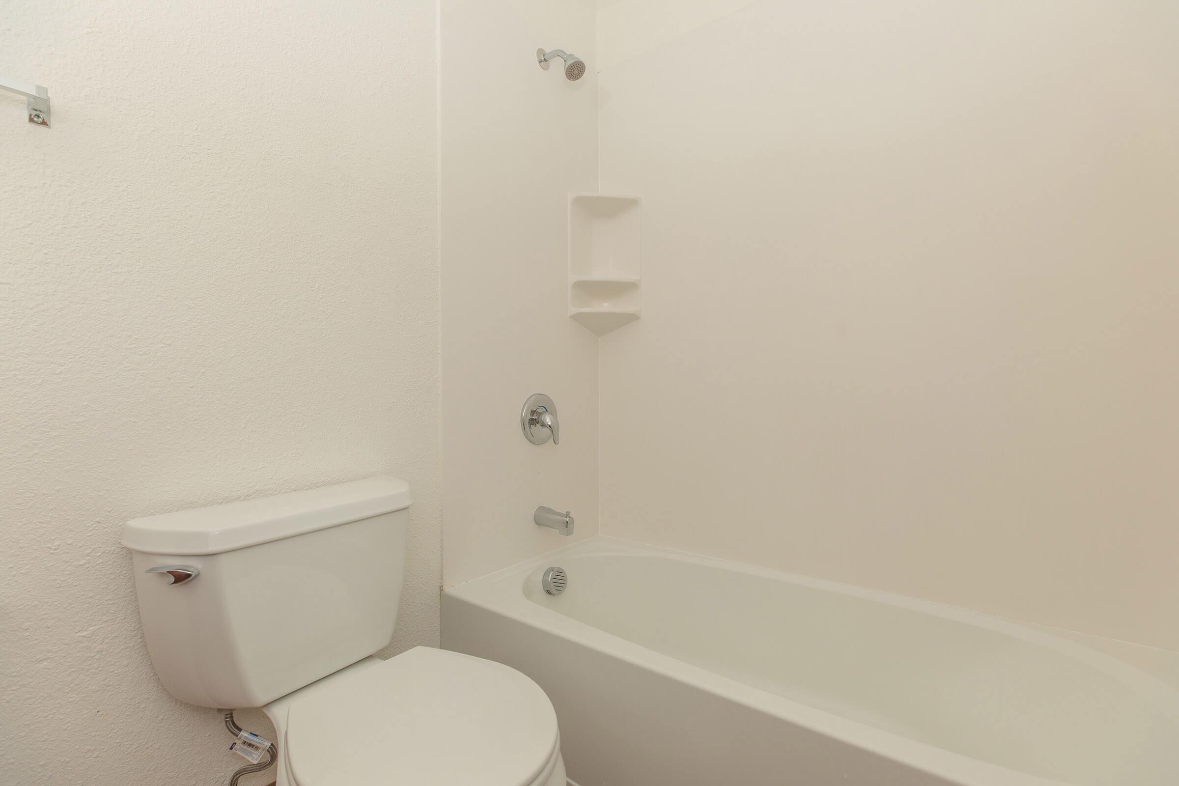 A simple bathroom featuring a white bathtub with a showerhead, a white toilet, and a minimalistic design with plain light-colored walls. There are built-in shelves in the wall beside the bathtub.