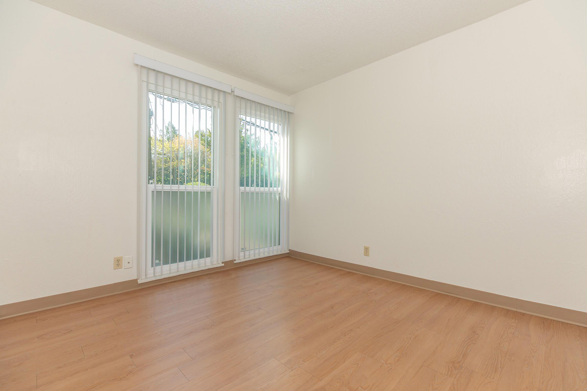A spacious, empty room with light-colored walls and wooden flooring. Two large windows with white blinds allow natural light to enter, providing a view of greenery outside. The room has a minimalistic design, creating a clean and airy atmosphere.