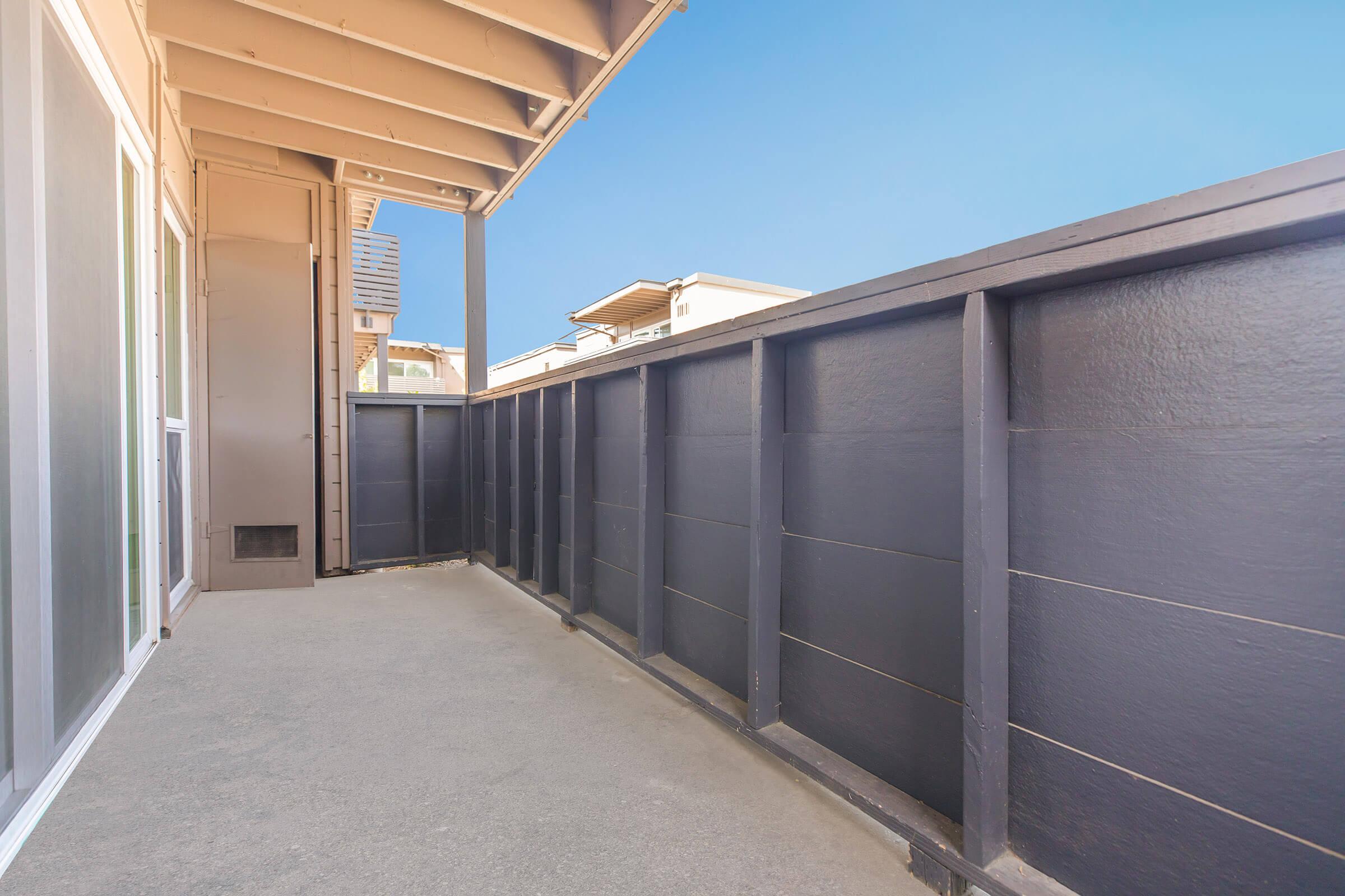 A balcony or patio view with a concrete floor and a dark wooden railing. The sky above is clear and blue, while the surrounding buildings are partially visible in the background. The space appears clean and well-maintained, offering a serene outdoor area.