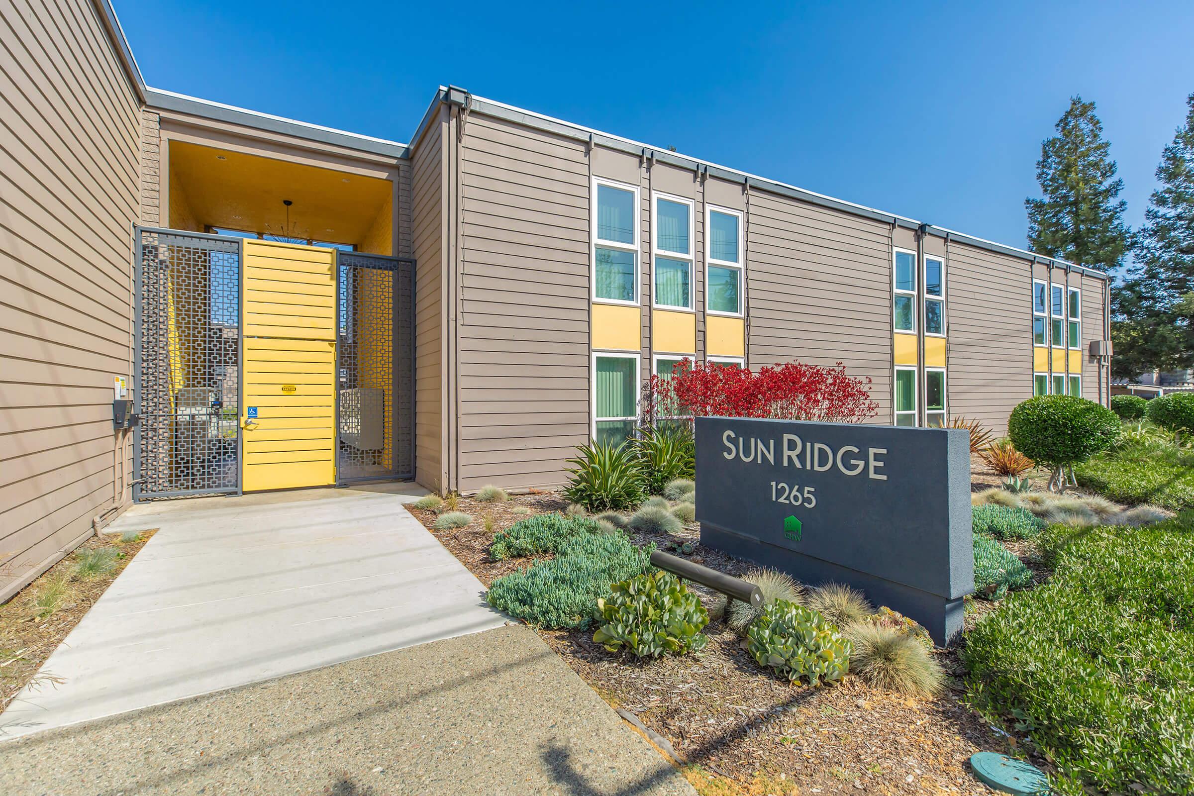 Front view of the Sun Ridge apartment complex, featuring a modern design with wooden siding and colorful accents. A welcoming entrance is marked by a yellow door and surrounded by well-maintained landscaping, including shrubs and decorative plants. The address, 1265, is displayed prominently on a sign.