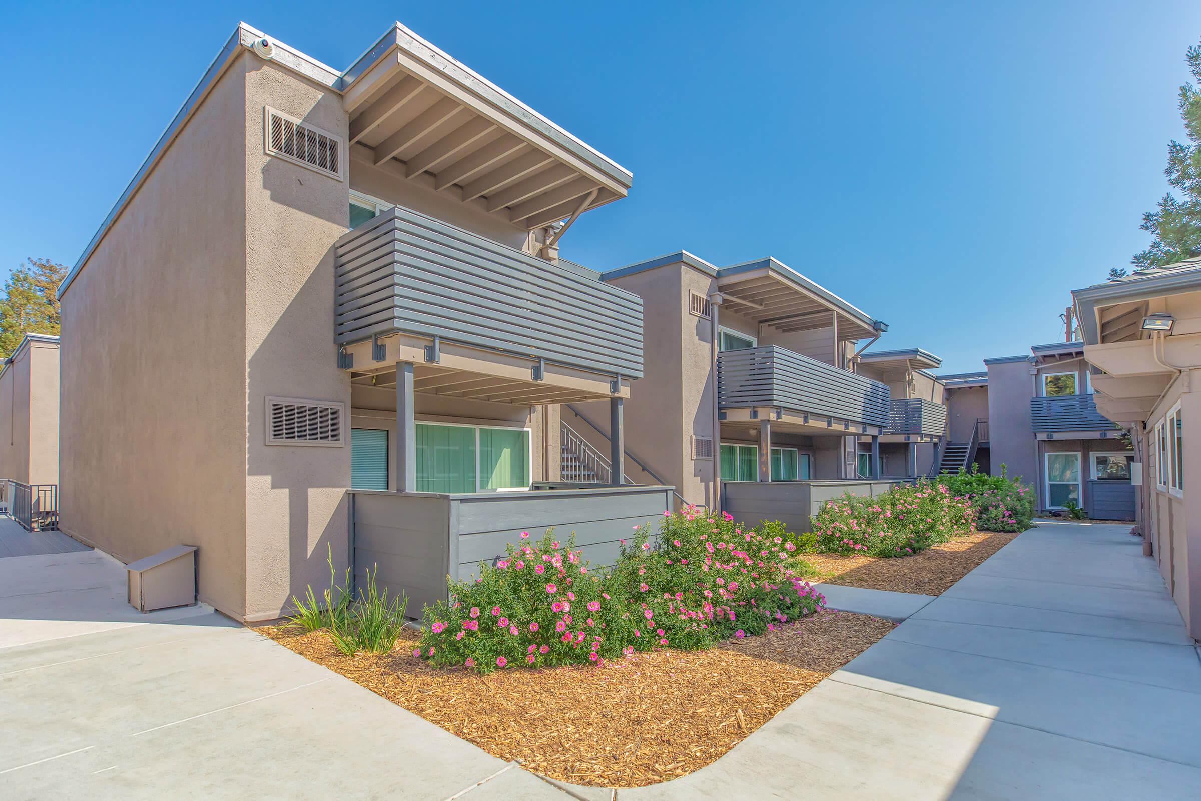 A modern apartment complex with two-story buildings featuring balconies. Flower beds with blooming pink roses line the pathway, providing greenery. The clear blue sky adds to the bright and inviting atmosphere. Pathways lead to the entrances of the buildings, which have a contemporary architectural design.