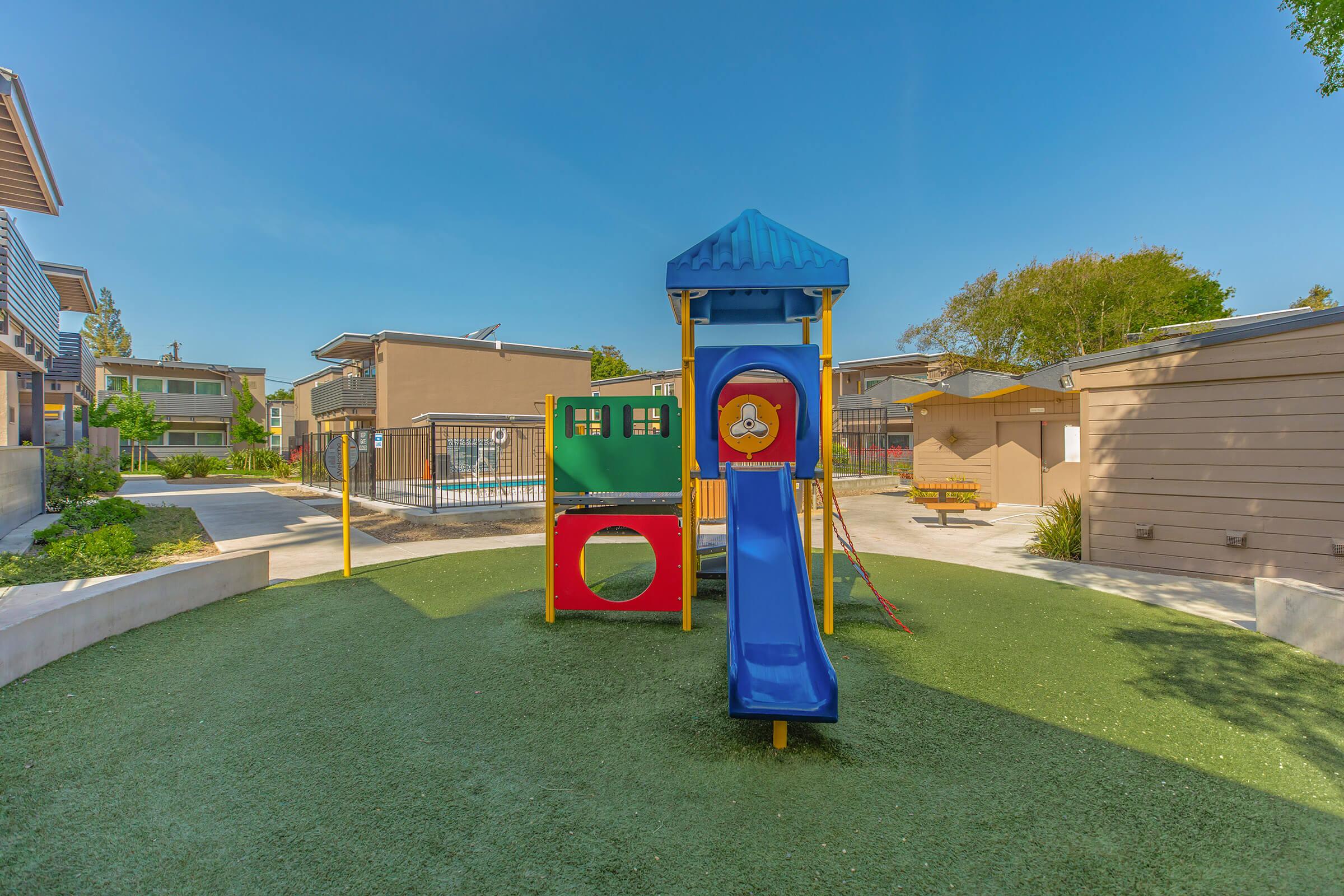 Colorful playground with a slide and climbing structure on green turf. In the background, there is a pool area and several residential buildings. The sky is clear and blue, and the environment appears bright and inviting.