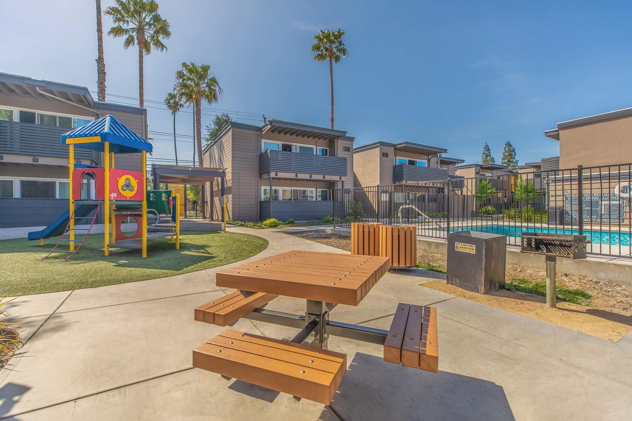 A sunny residential area featuring a playground with a slide and climbing structure, a picnic table, and a swimming pool surrounded by palm trees. Modern buildings are in the background, creating a vibrant, family-friendly environment.