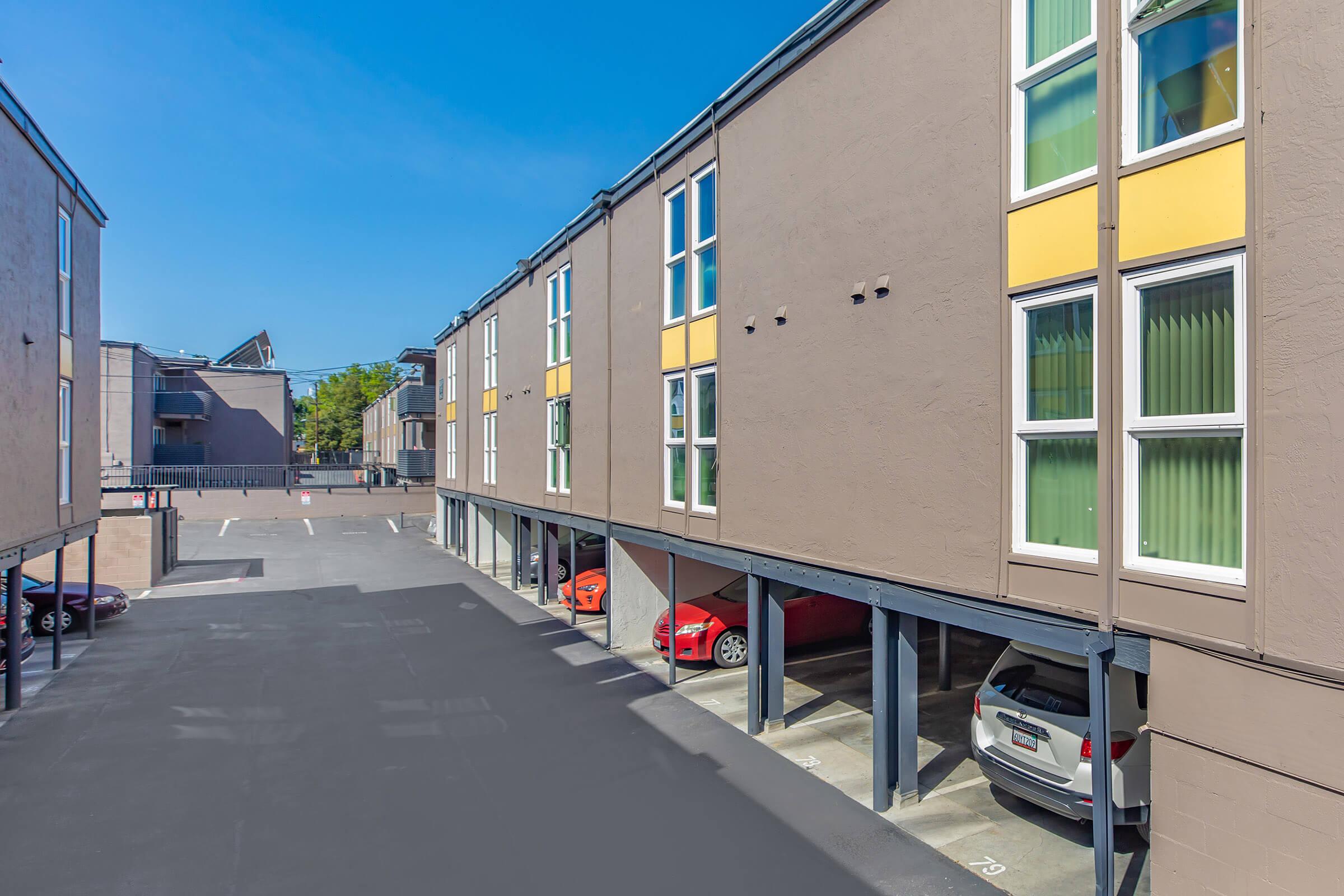 View of an apartment complex's exterior featuring two buildings with several parking spots underneath. Several cars are parked in the covered spaces, and the sky is clear and blue. The buildings have modern designs with large windows and a mix of beige and yellow colors.