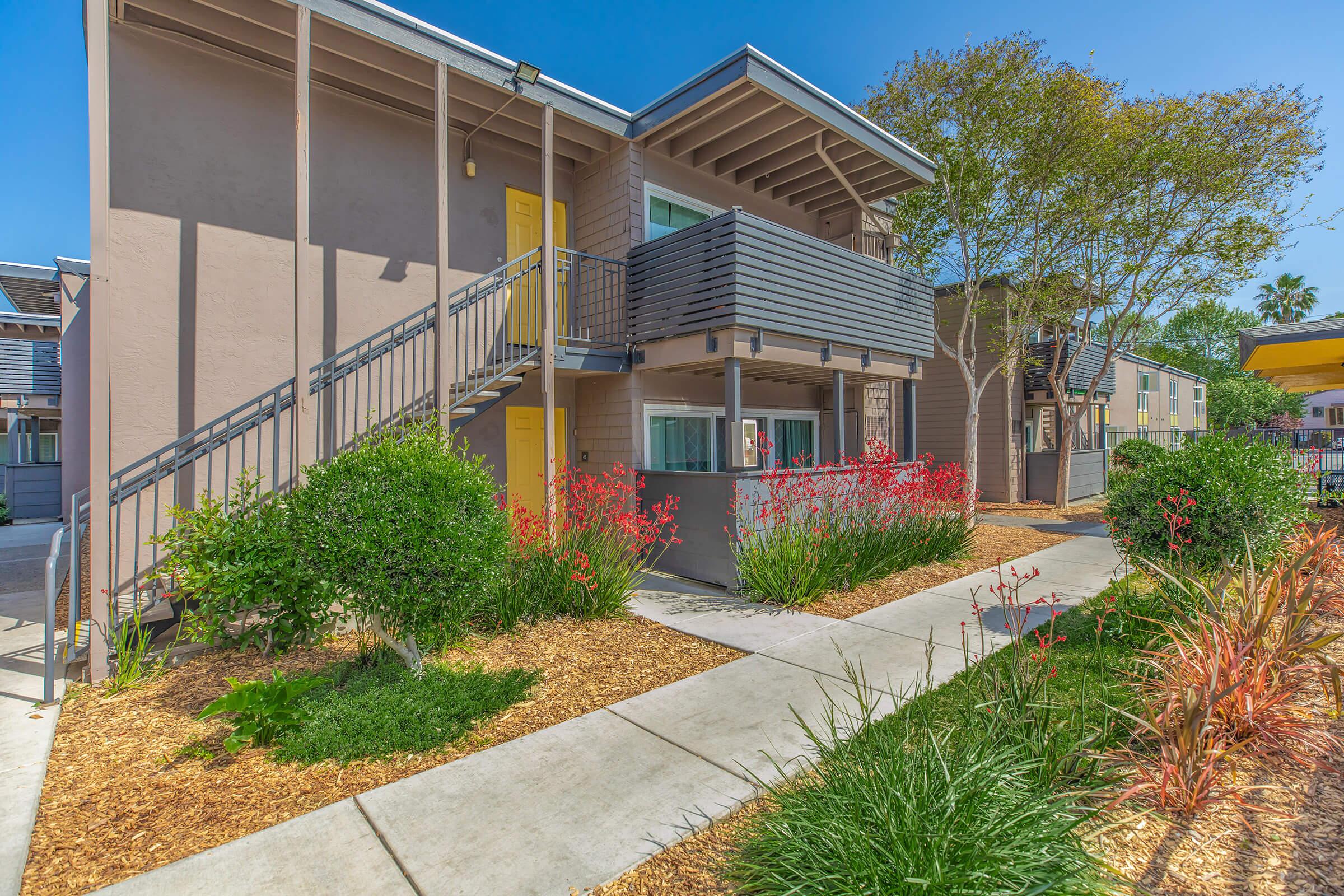 A landscaped courtyard featuring a modern, multi-story apartment building with yellow doors. The pathway is lined with colorful plants and shrubs, and there are trees providing shade. Bright blue skies above add to the inviting atmosphere.