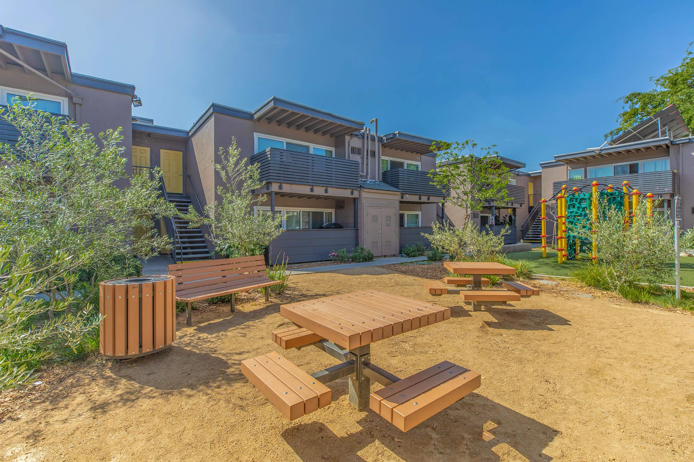 Outdoor area featuring several wooden picnic tables surrounded by shrubbery and landscaping. In the background, there are modern buildings with balconies, and to the side, a colorful playground structure. The sky is clear and blue, creating a bright and inviting atmosphere.