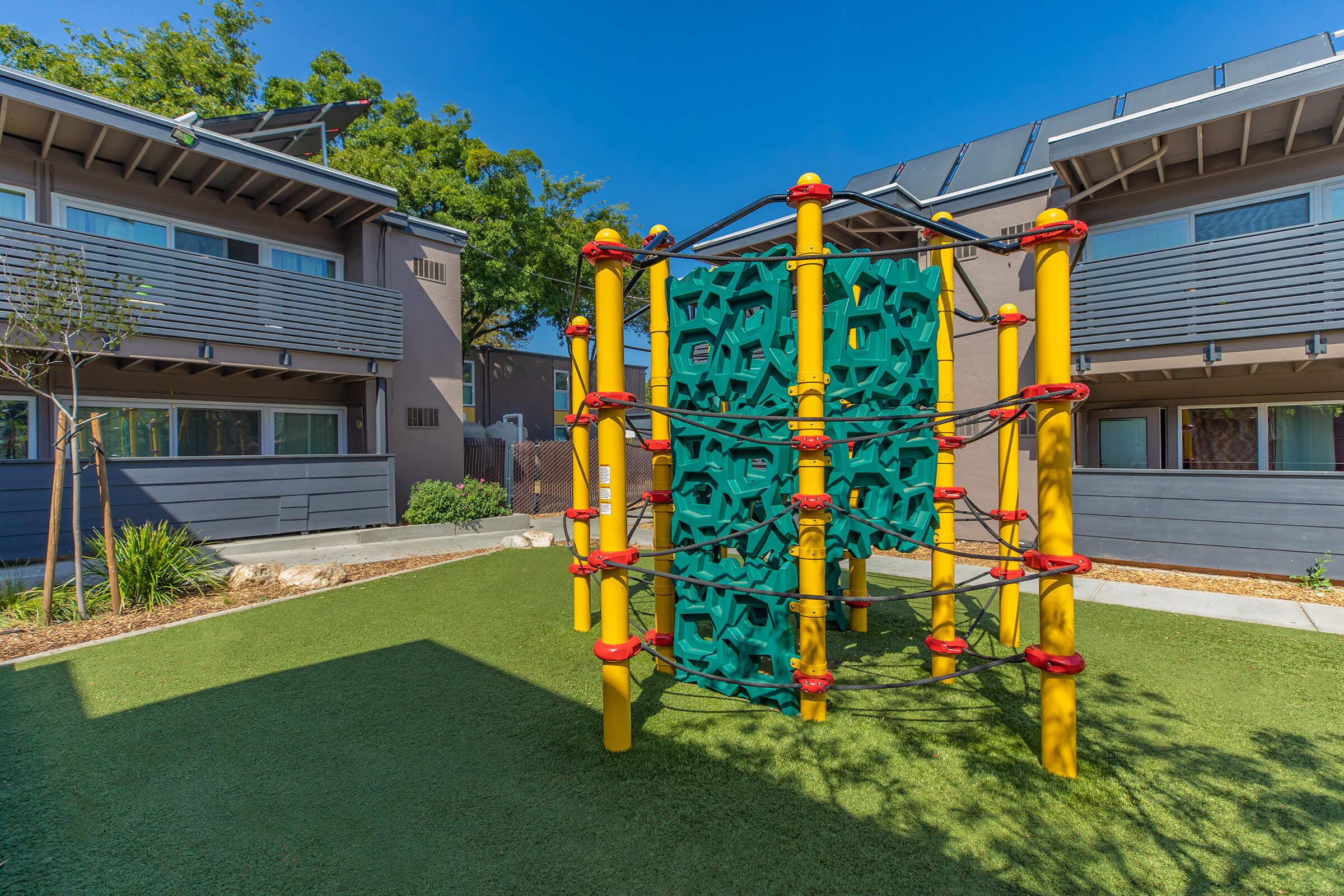 A colorful children's climbing structure made of green and yellow materials is situated in a grassy play area, surrounded by modern buildings with grey exteriors and bright blue skies overhead. The setting appears inviting and safe for kids to play.