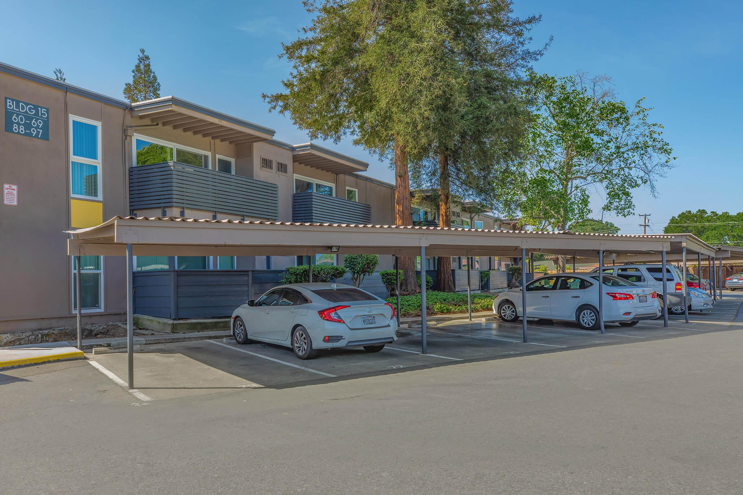 A parking lot with several cars parked under covered spaces, adjacent to a residential building with multiple apartments. The building features balconies and greenery, with a clear blue sky in the background.