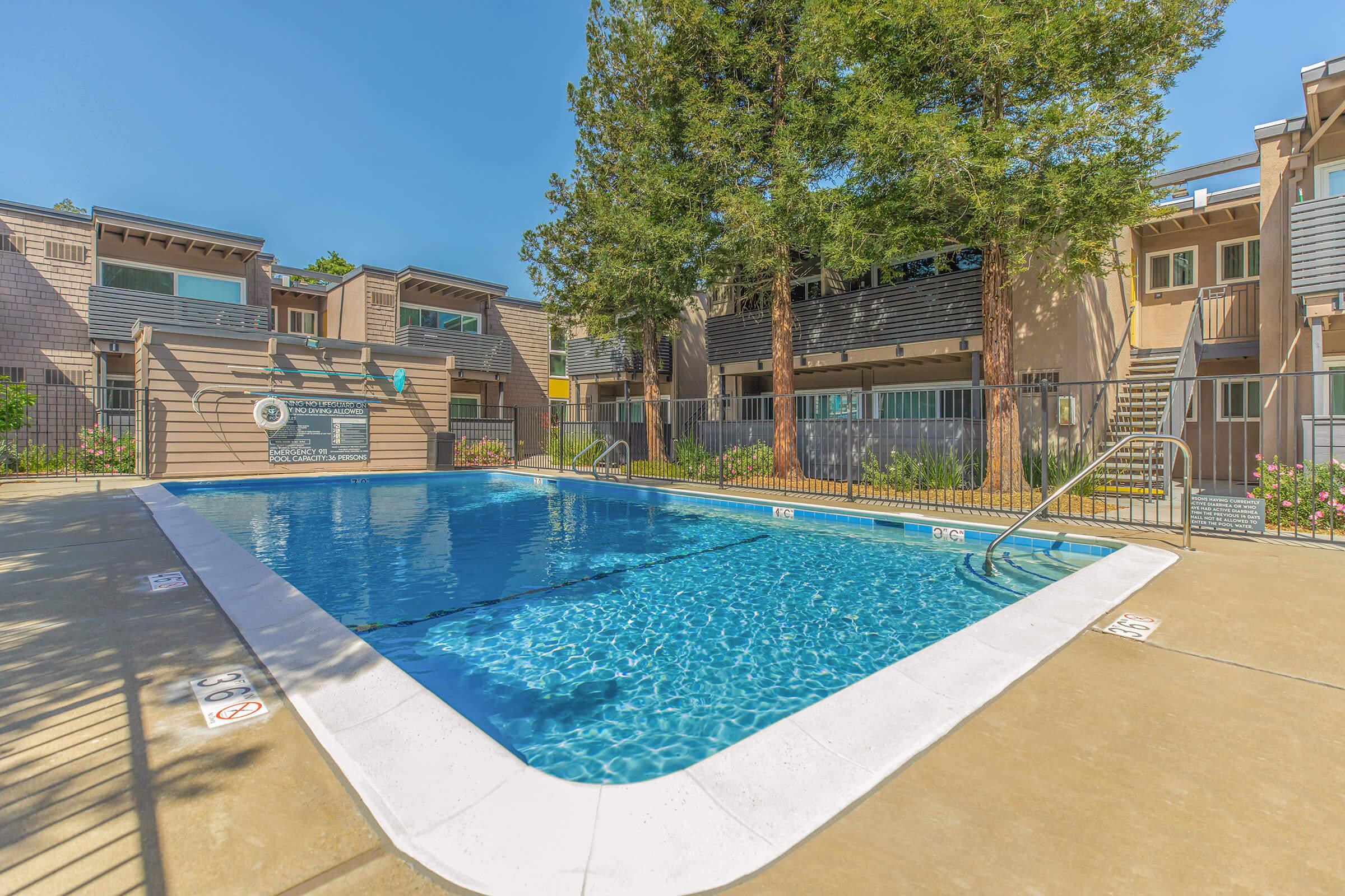 A clear blue swimming pool surrounded by a concrete deck, featuring a railing and steps leading into the water. Lush green trees and multi-story apartment buildings are visible in the background under a bright blue sky. The scene conveys a relaxing outdoor environment ideal for leisure.