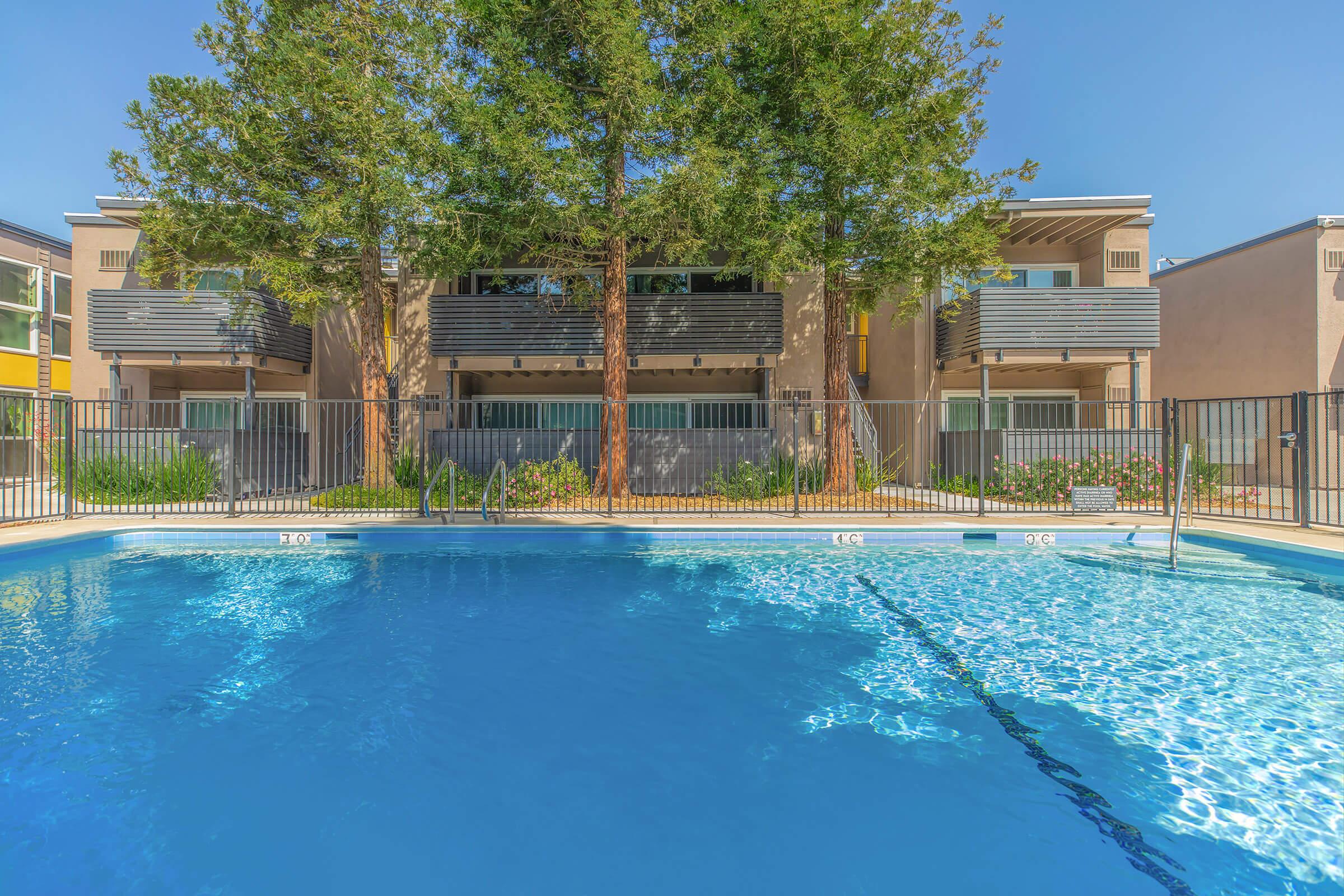 A clear blue swimming pool with steps leading into it, surrounded by a fence. In the background, there are modern apartment buildings featuring balconies, greenery, and trees under a clear blue sky. The scene conveys a relaxing, sunny atmosphere.