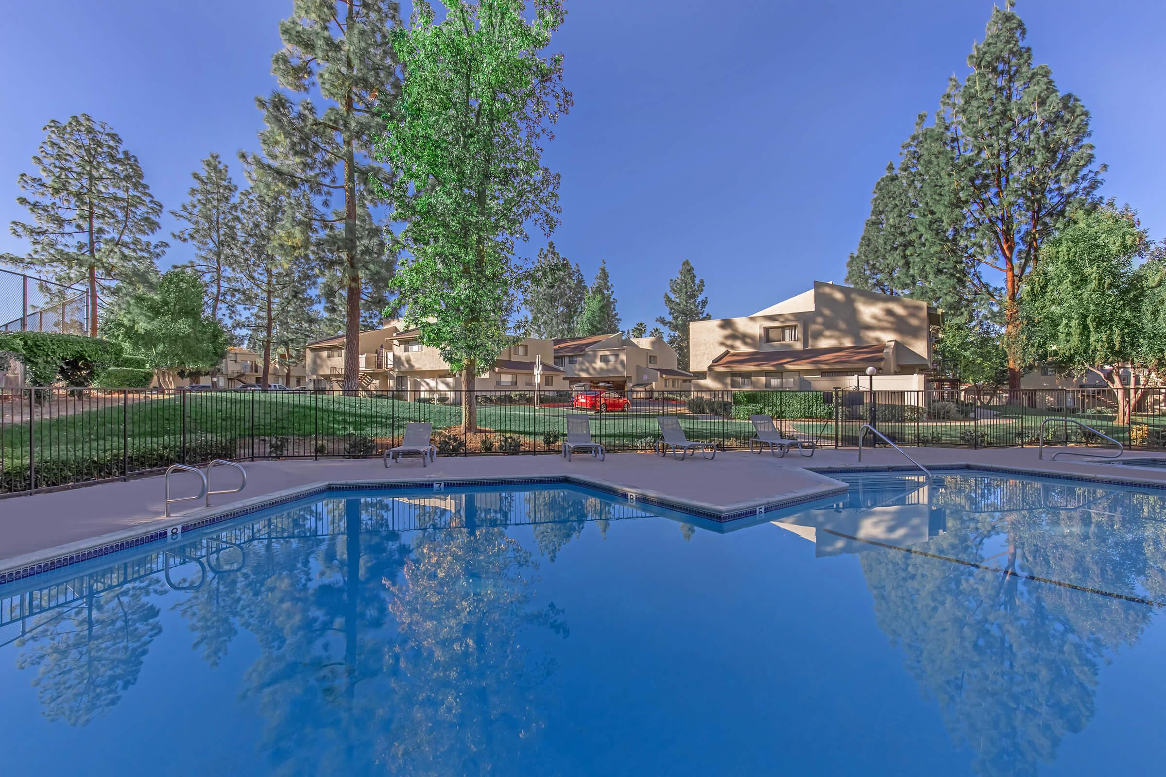 A serene pool area surrounded by lush trees and greenery. The calm water reflects the blue sky and nearby residential buildings. Sunlight filters through the trees, creating a relaxing outdoor atmosphere perfect for leisure and relaxation. The scene conveys a peaceful suburban environment.