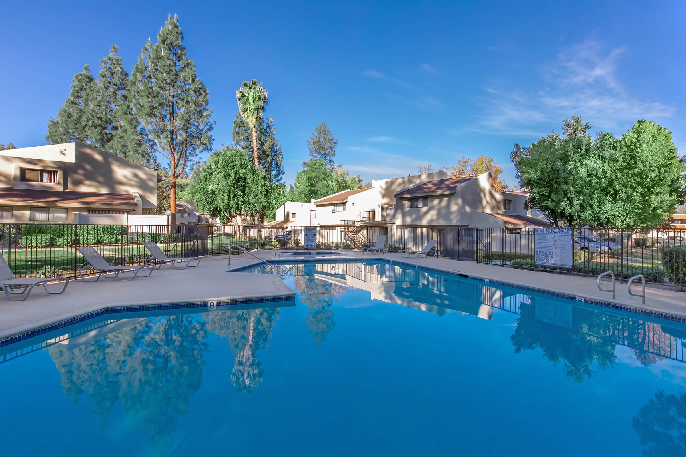 A clear swimming pool surrounded by lounge chairs and lush greenery. Residential buildings with light-colored exteriors line the pool area under a blue sky with a few clouds. Trees provide shade, creating a tranquil atmosphere.