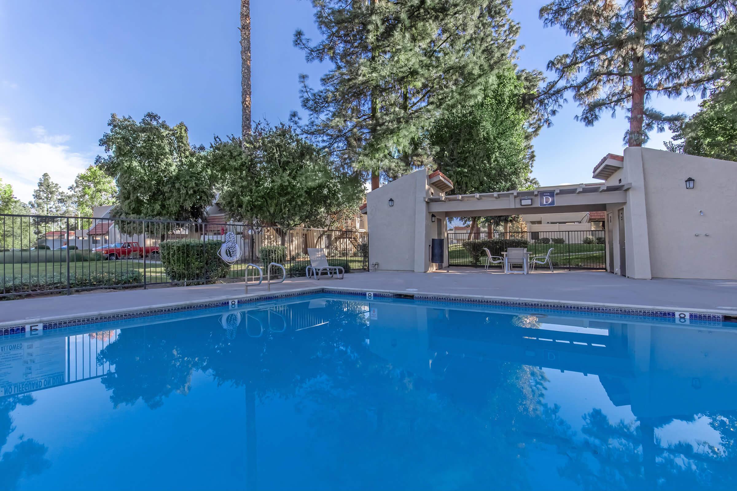 A clear blue swimming pool surrounded by green trees and a well-maintained landscape. There are lounge chairs by the poolside and a shaded seating area under a gazebo. The sky is bright with a few clouds, creating a serene and inviting atmosphere.