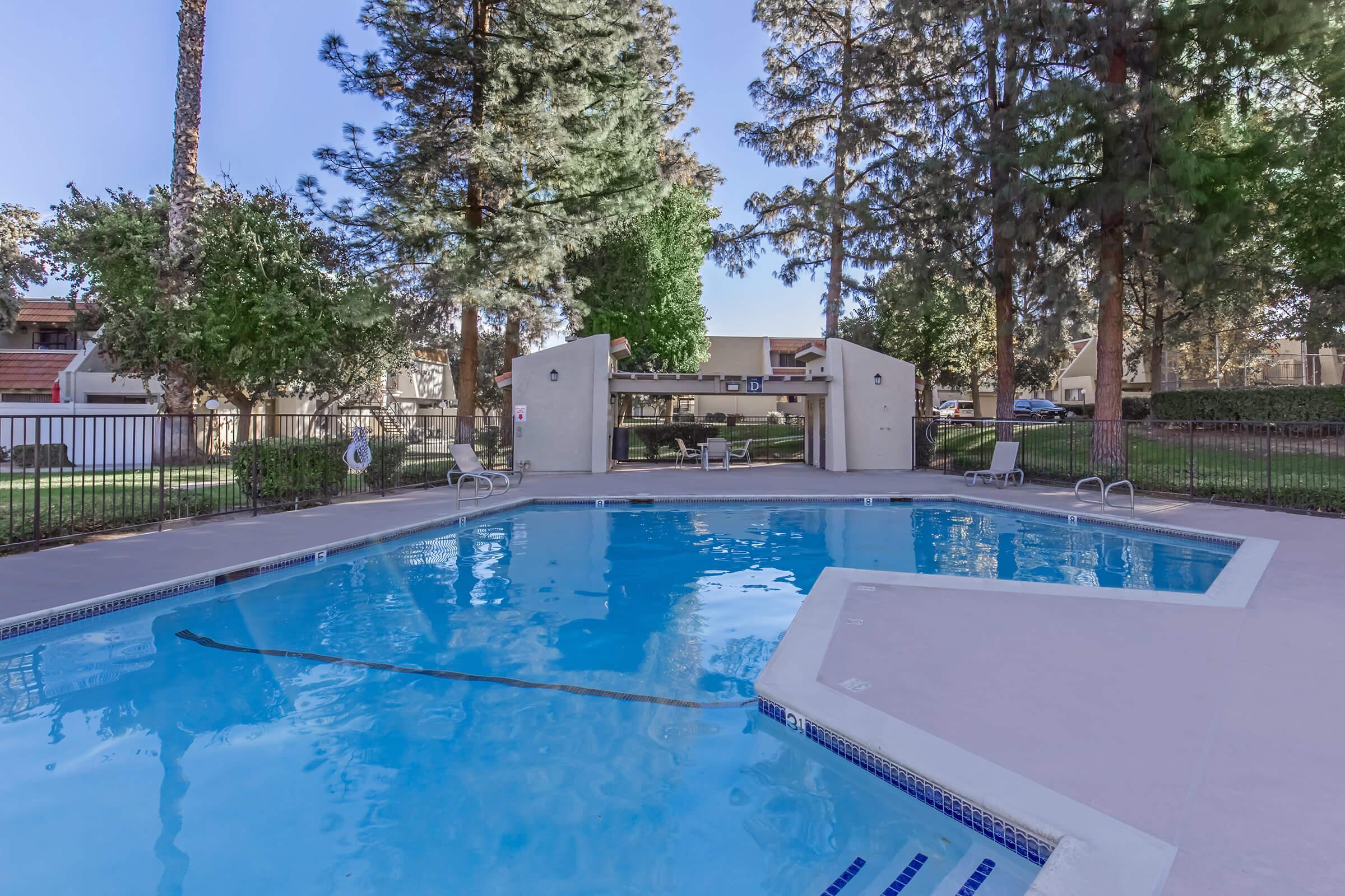 A clear blue swimming pool surrounded by tall trees and greenery. The pool area features lounge chairs and a fenced perimeter. In the background, there are buildings that suggest a residential complex. The scene is bright and inviting, ideal for relaxation or recreation.