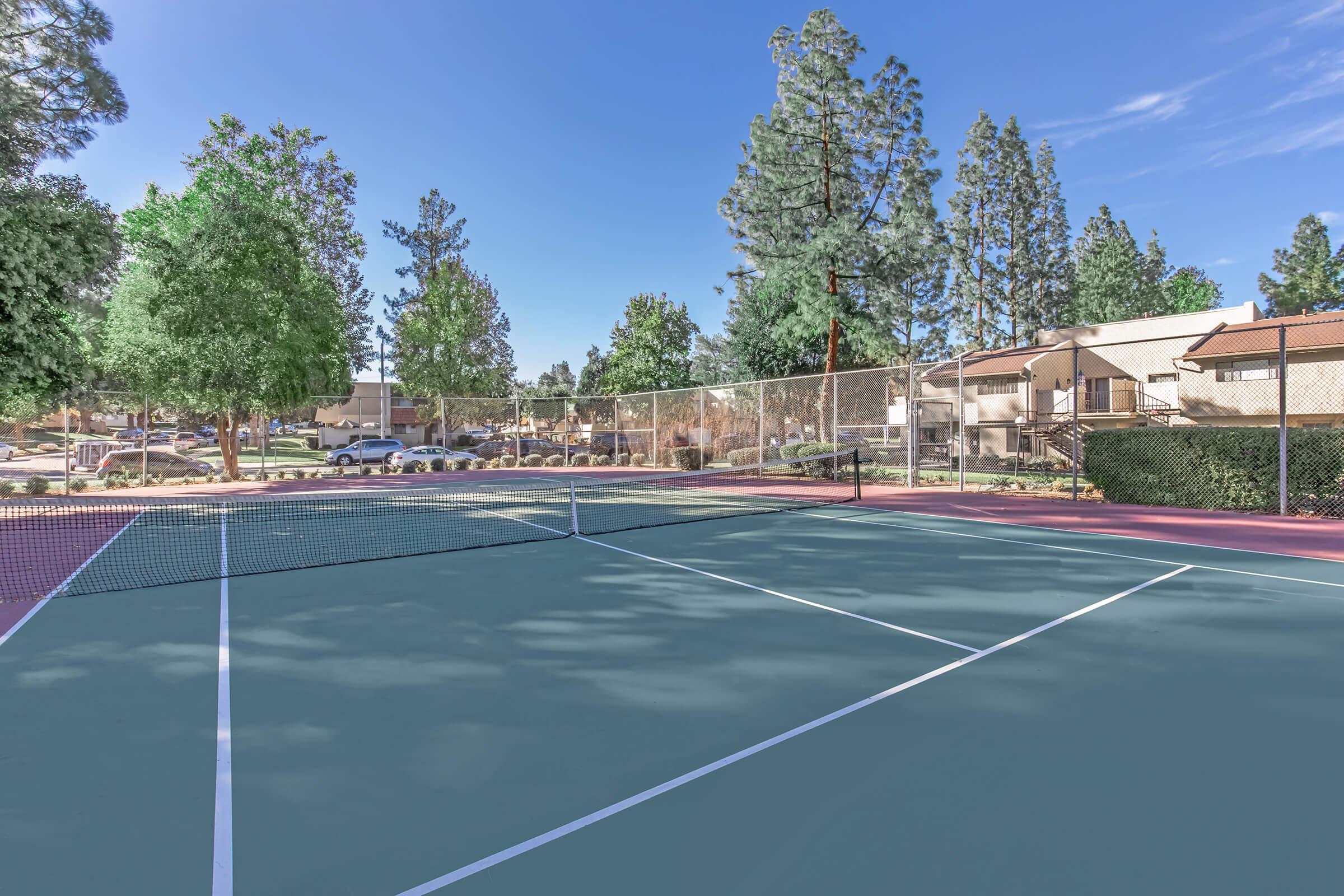 A well-maintained outdoor tennis court surrounded by tall trees and greenery, with residential buildings visible in the background. The court features a net in the center and lines clearly marked on the surface under a clear blue sky.