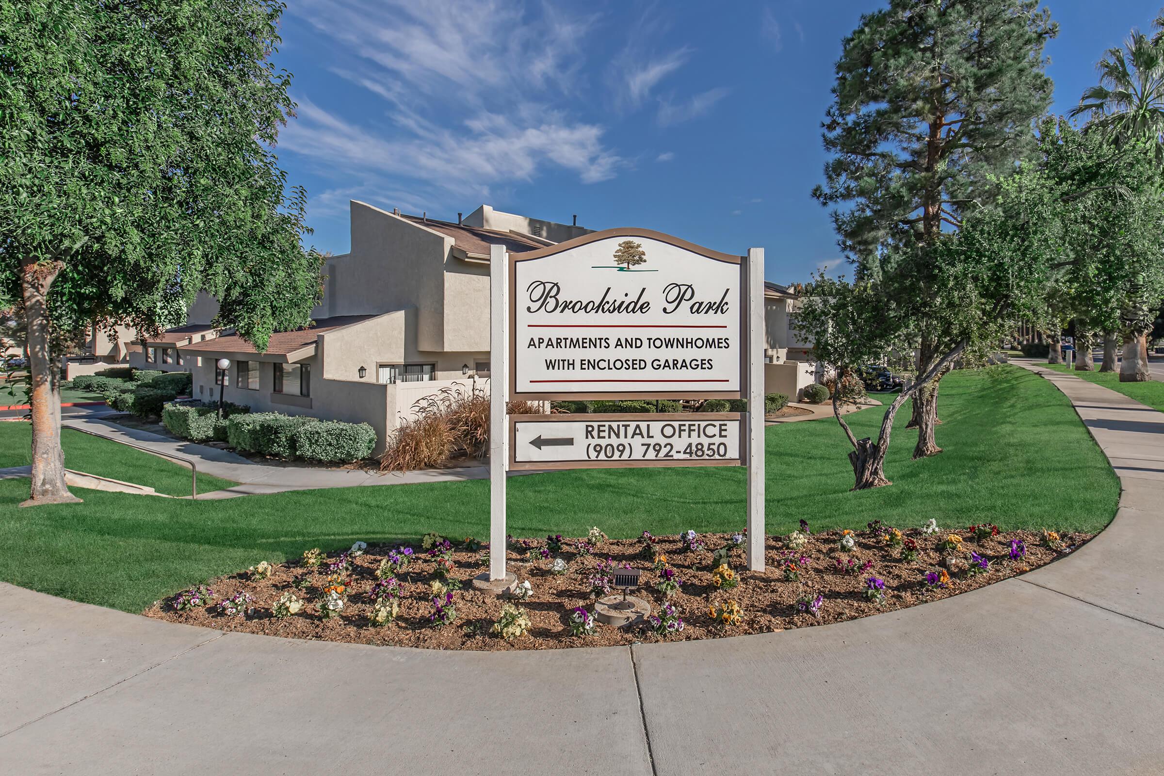 Sign for Brookside Park, showcasing apartments and townhomes with enclosed garages. Includes a direction arrow pointing to the rental office with the phone number (909) 792-4850. The background features green lawns, trees, and a clear blue sky.