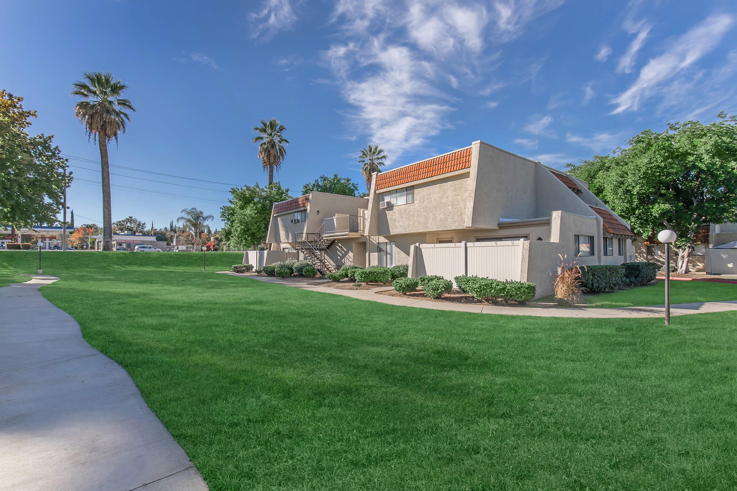 A well-maintained residential area featuring modern apartment buildings surrounded by lush green lawns and palm trees under a clear blue sky. The walkway leads through the manicured landscape, creating an inviting atmosphere.