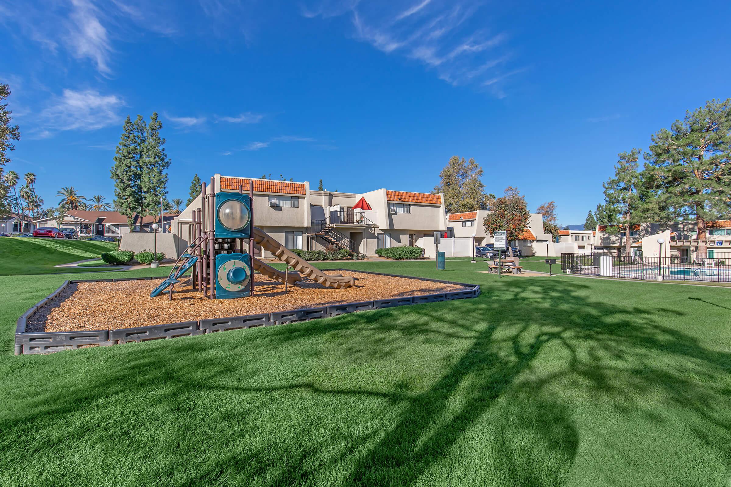 Playground with a slide and climbing structures on a sunny day. Lush green grass surrounds the play area, and there are trees in the background. Several residential buildings are visible in the distance under a clear blue sky.