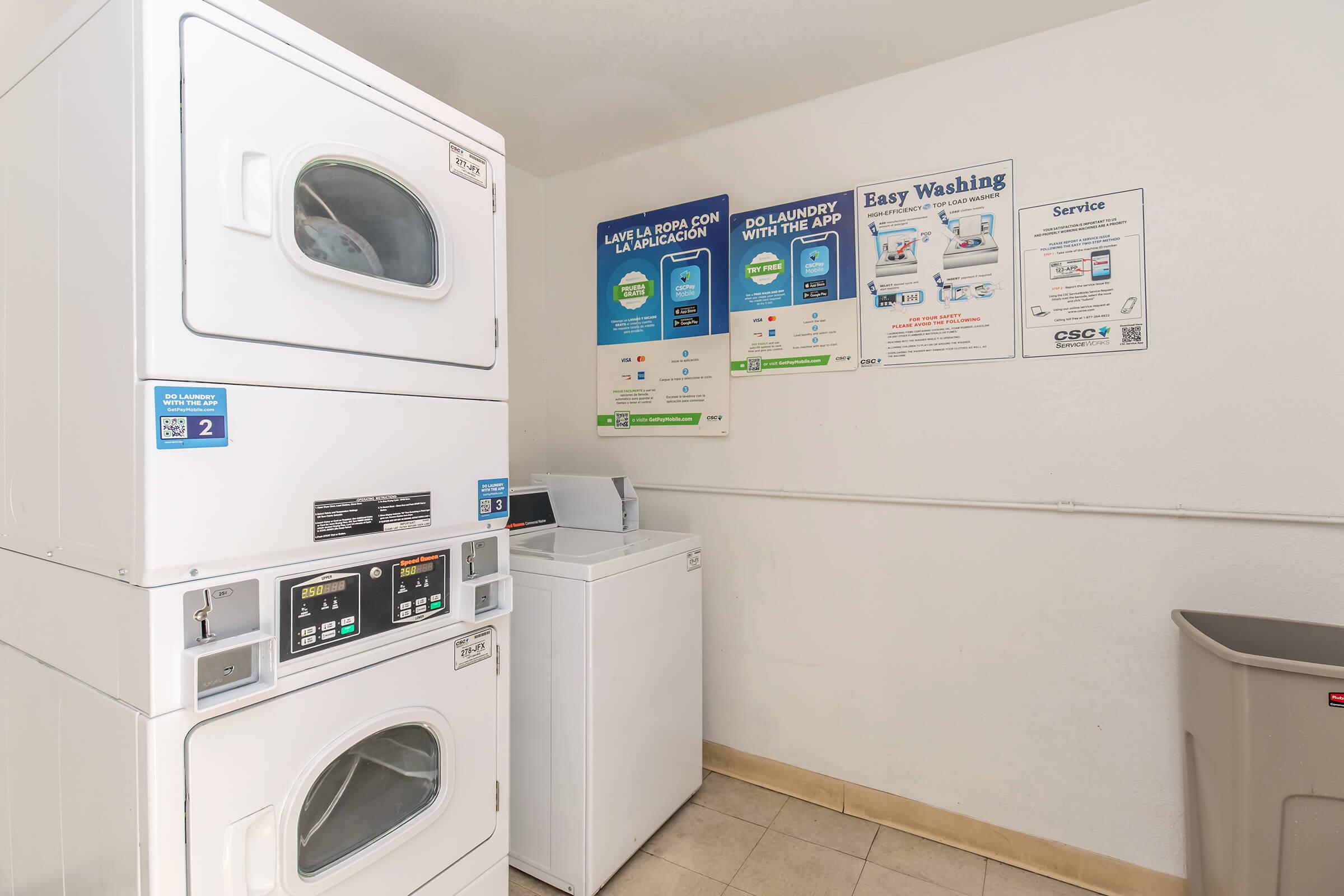 A laundry room featuring a stacked washer and dryer unit alongside a single washing machine. There are informational signs on the wall regarding laundry services and payment options. A trash bin is located in the corner of the room. The flooring is tiled and the walls are plain.