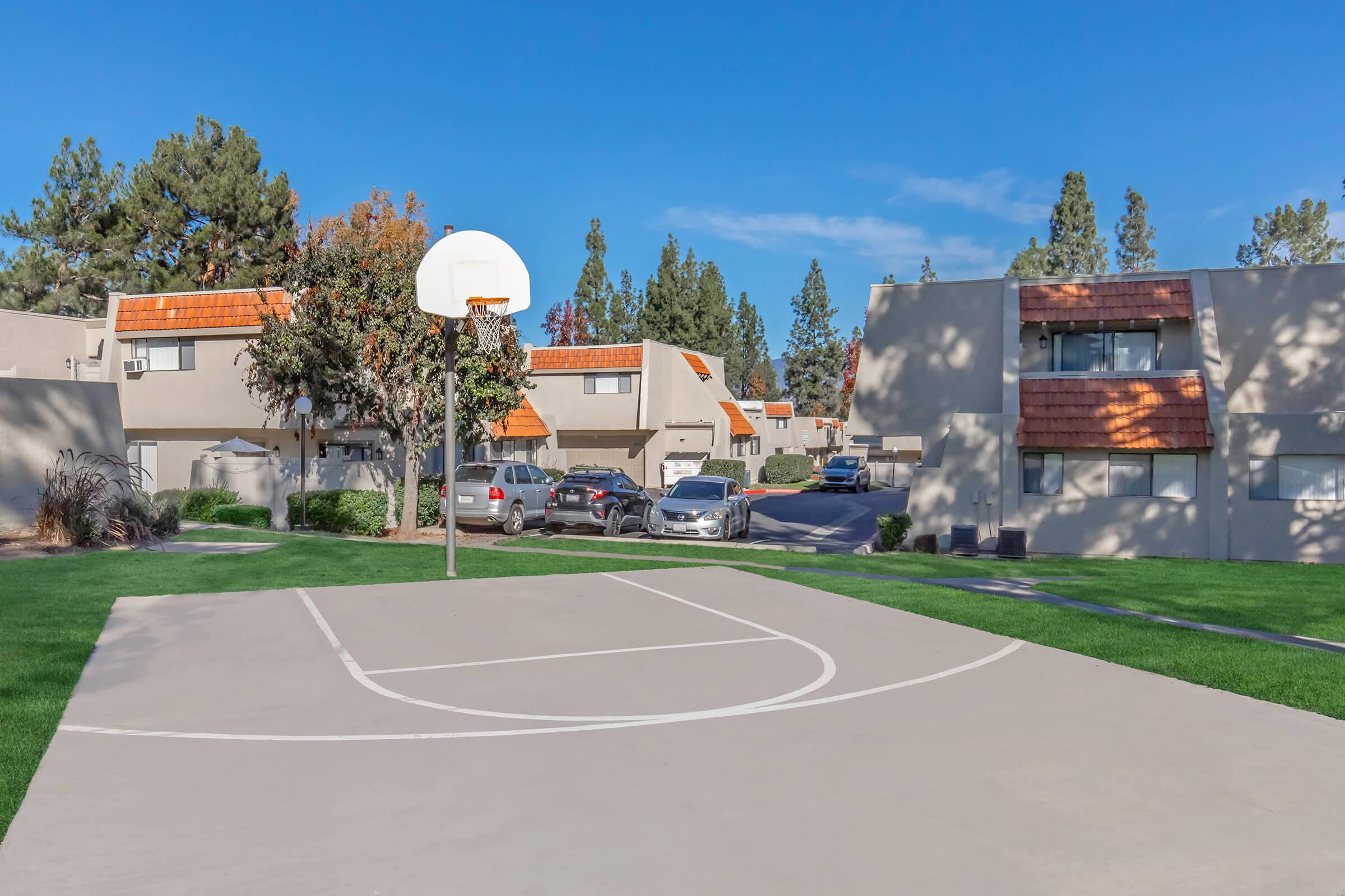A basketball court with a hoop in the foreground, set within a residential area. Apartment buildings with orange-tiled roofs and green lawns are visible in the background, surrounded by trees under a clear blue sky. Vehicles are parked nearby, and the scene is well-lit and inviting.