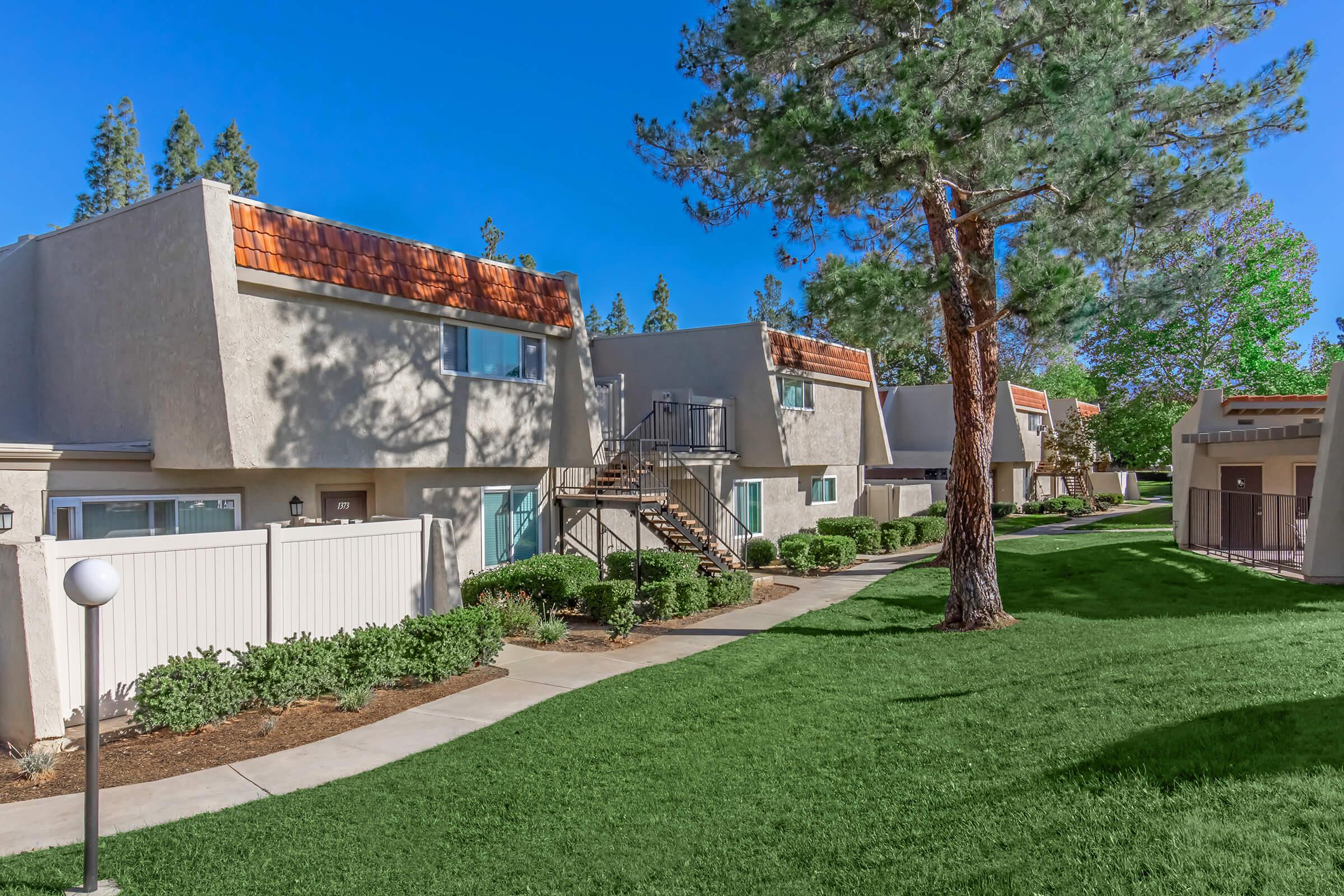 A well-maintained residential complex featuring modern townhouses with orange roofs, surrounded by lush green grass and neatly trimmed shrubs. A pathway leads through the property, and a tall tree adds shade to the area, creating a pleasant and inviting atmosphere for residents.
