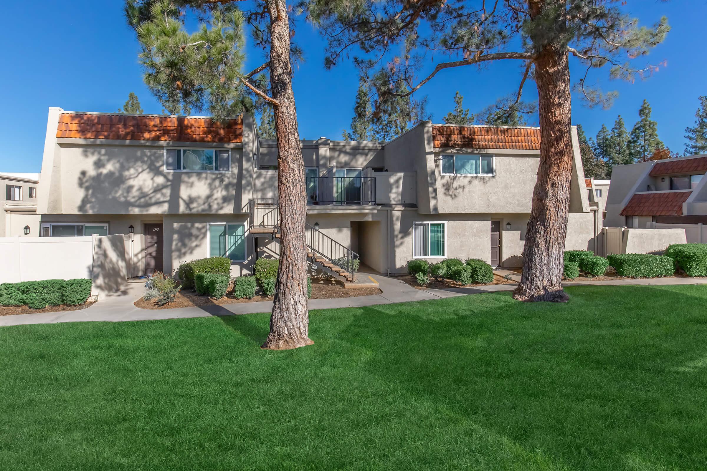Two-story apartment buildings with a walkway lined by green lawns and shrubs. Trees are in the foreground, and the sky is clear and blue. The architecture features a combination of light-colored walls and reddish roof tiles.