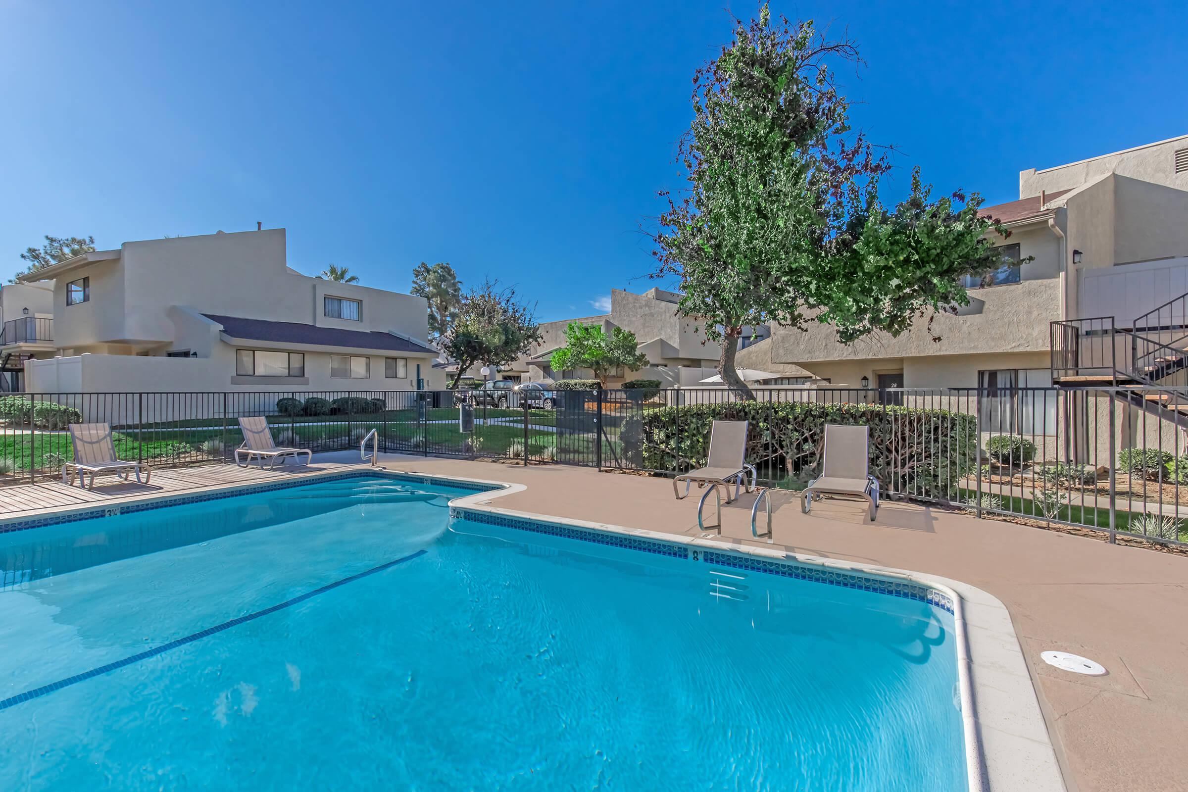 A clear blue swimming pool surrounded by lounge chairs and a fenced area. In the background, there are residential buildings and green trees under a bright blue sky. The scene is inviting and suggests a relaxed outdoor atmosphere, perfect for leisure and enjoyment.