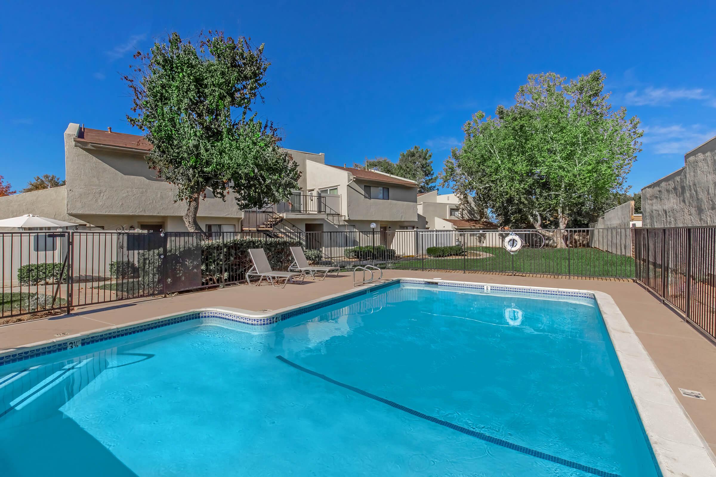 A clear blue swimming pool surrounded by a fence, with poolside lounge chairs. In the background, there are multi-storied residential buildings and lush trees under a bright blue sky. The scene conveys a serene and inviting atmosphere.