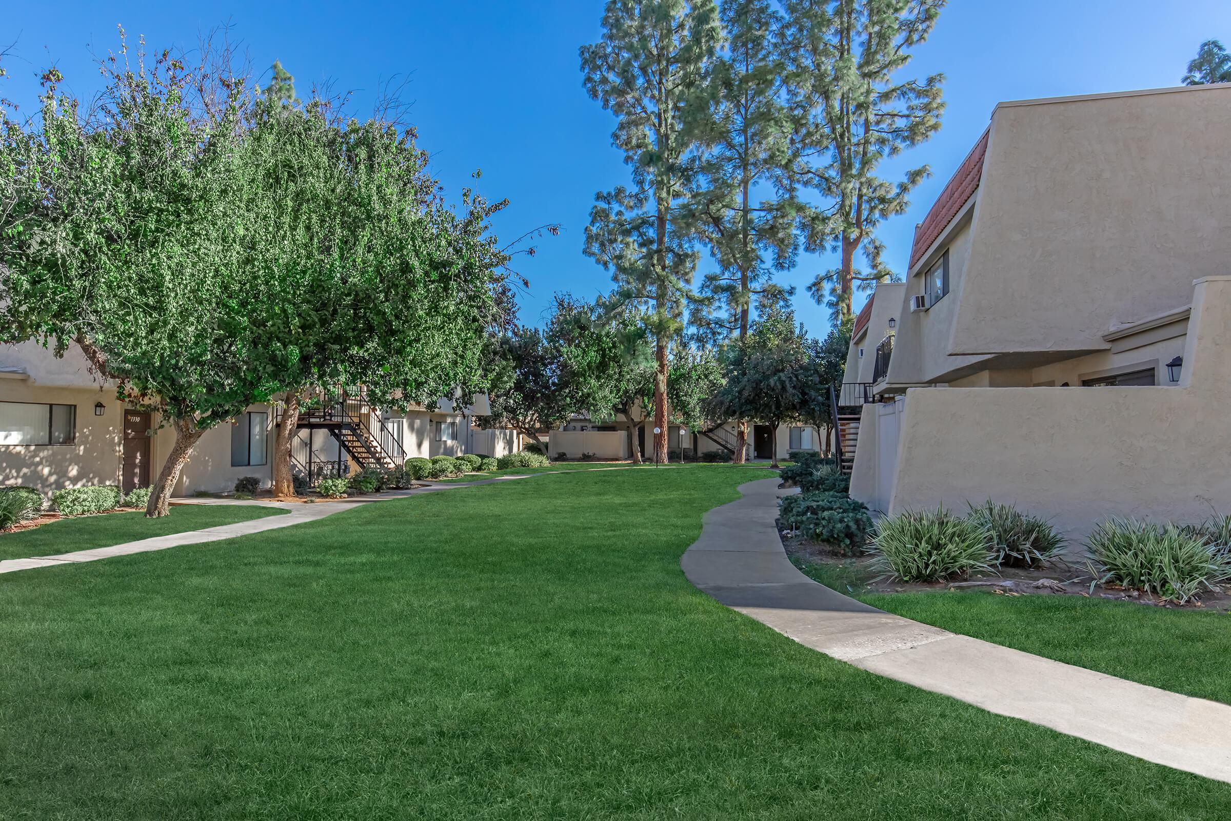 A peaceful residential area featuring well-maintained green lawns and pathways. The scene includes tall trees, small shrubs, and buildings arranged around a central lawn, under a clear blue sky. The setting is inviting and serene, perfect for walks or relaxation.