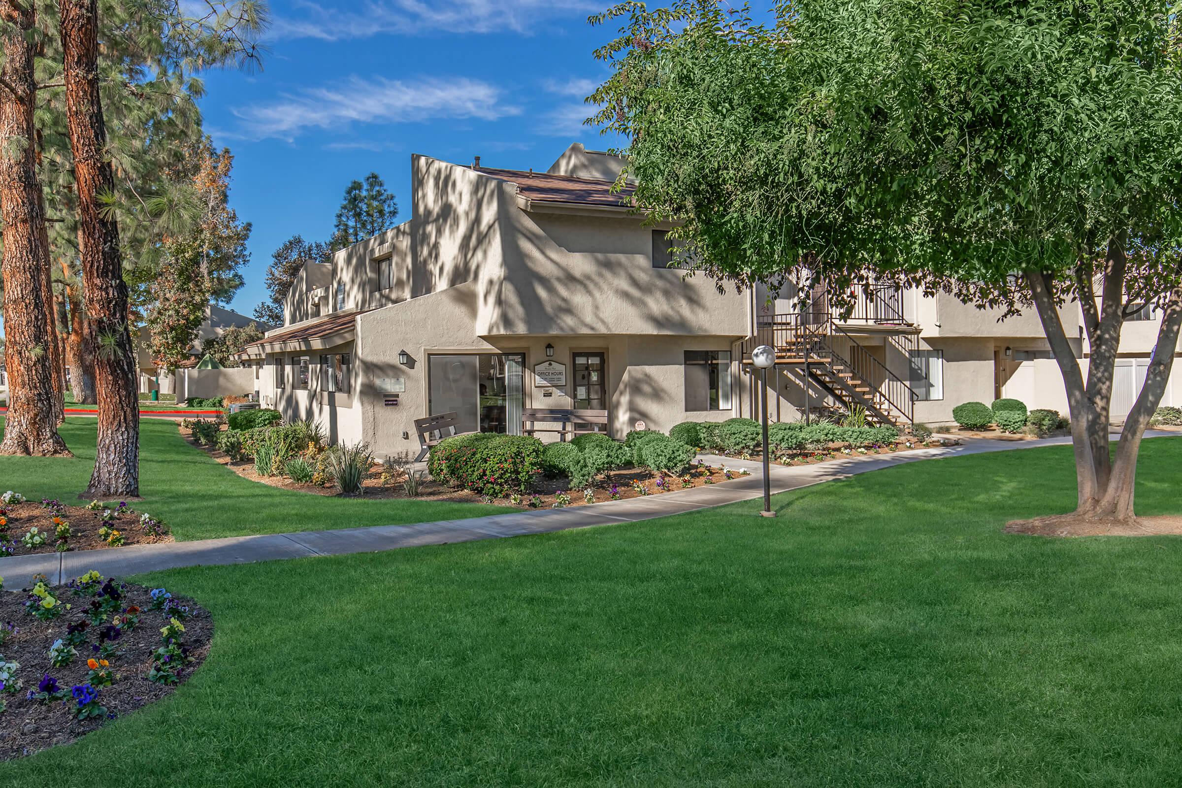 A landscaped area featuring a pathway lined with green grass, shrubs, and flowering plants. In the background, there are two-story apartment buildings with balconies, along with tall trees providing shade. The scene is sunny and well-maintained, creating an inviting outdoor space.