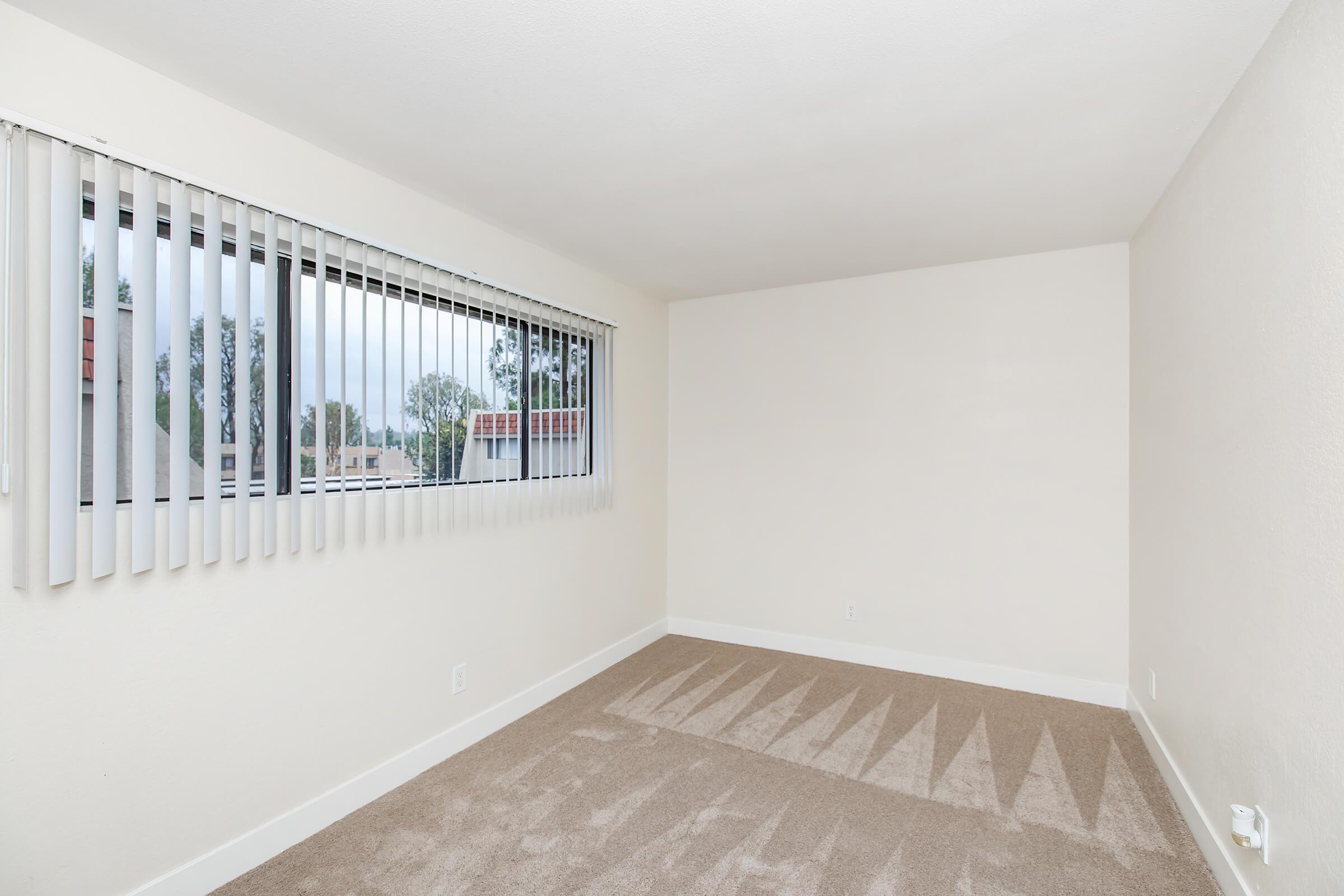 A vacant room with beige walls and light brown carpet. It features a large window covered with vertical blinds, allowing natural light to enter. The room has a simple and uncluttered appearance, with no furnishings or decorations, and visible shadows cast on the carpet from the window.