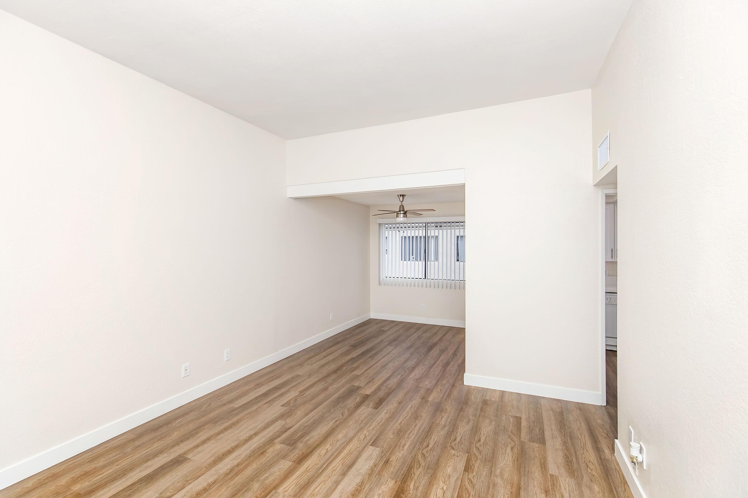 A spacious, empty room with light-colored walls and wooden flooring. There is a ceiling fan in the center and a window with blinds on the far wall, allowing natural light to illuminate the space. The design is minimalist and clean, making it ready for various furniture arrangements.