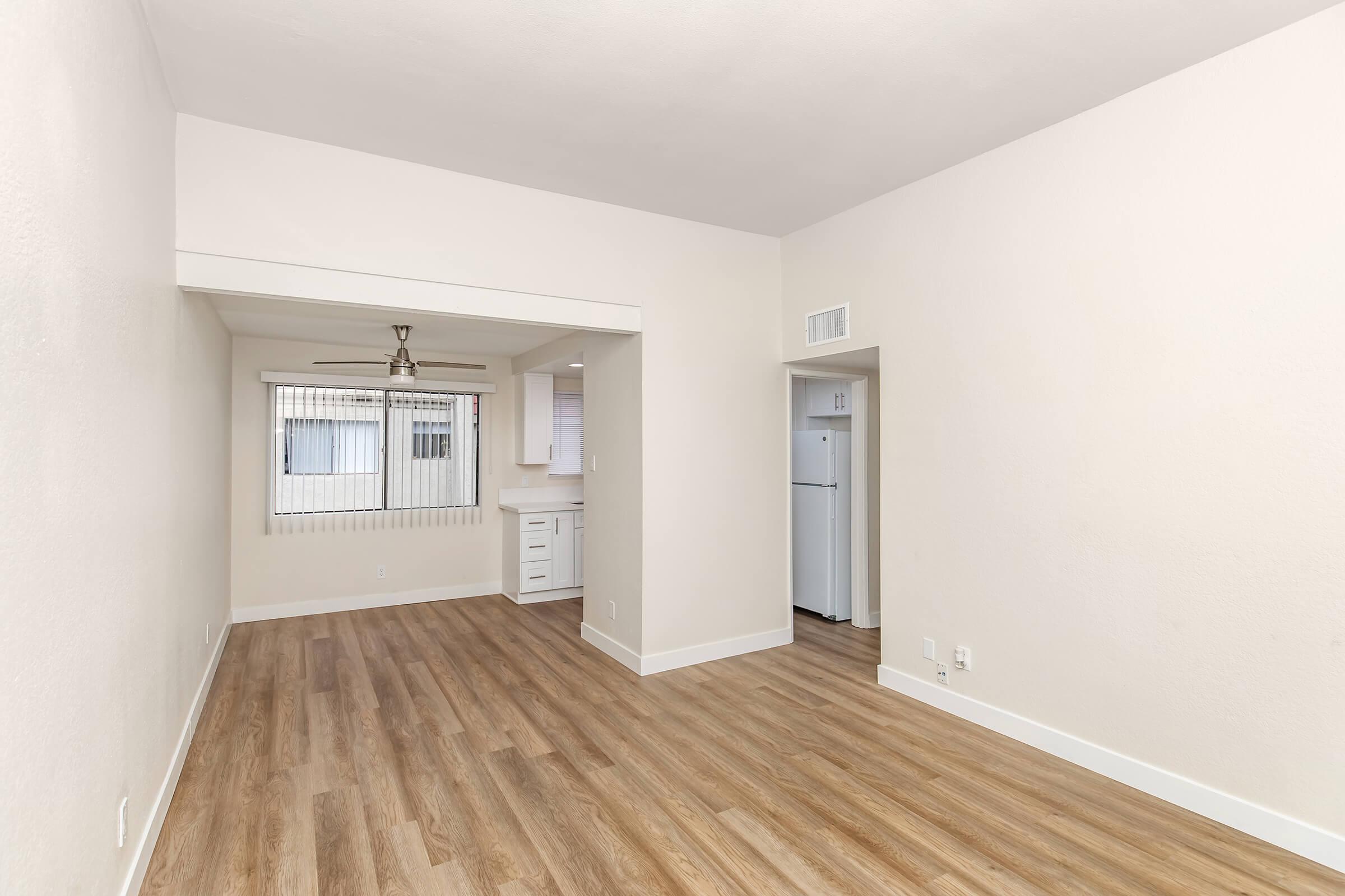 A bright, empty living space with light-colored walls and wooden flooring. There is a ceiling fan and a doorway leading to a kitchen area featuring a refrigerator. A window with vertical blinds allows natural light into the room, enhancing the spacious feel.