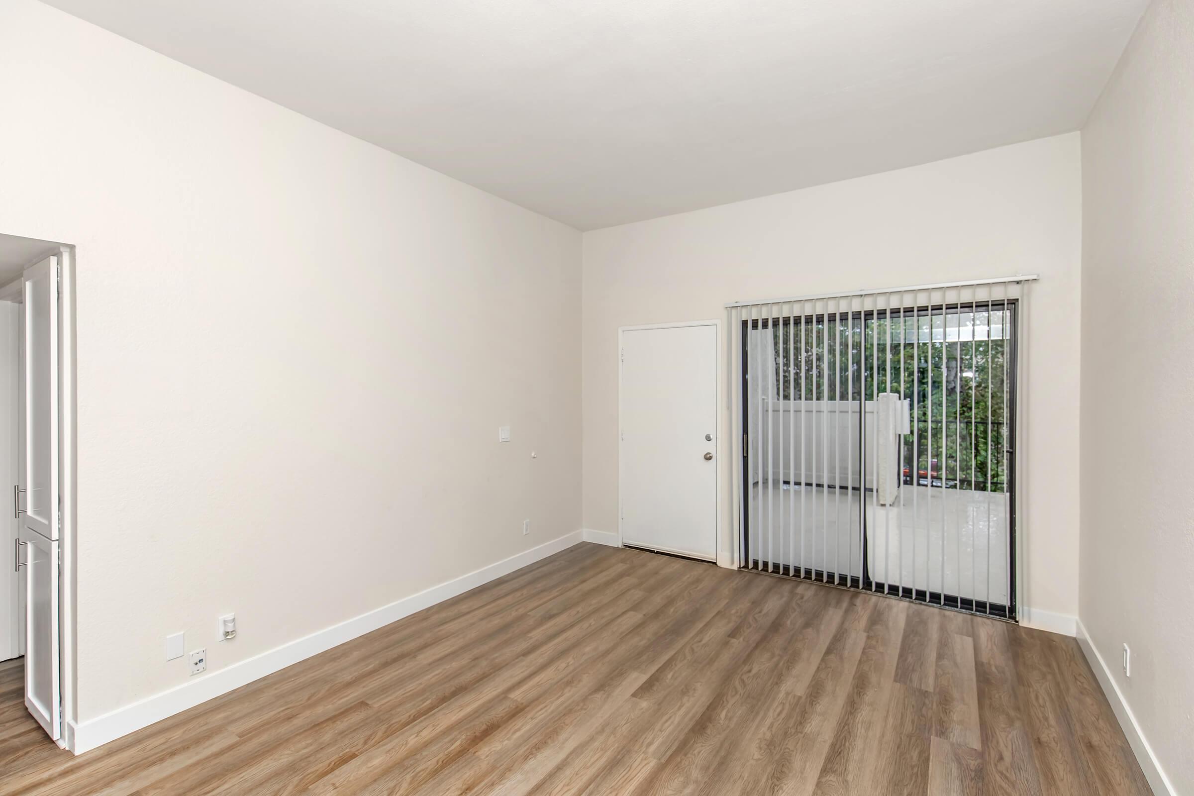 Empty room with light-colored walls and wooden flooring. A large sliding glass door with vertical blinds leads to an outdoor space, and there is a simple door on one wall. The room is bright with natural light coming through the door.