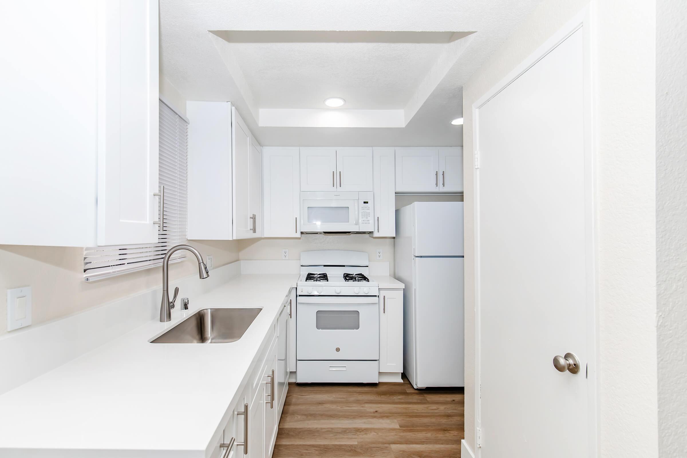A modern kitchen featuring white cabinetry, a stainless steel sink, a gas stove, a microwave, and a refrigerator. The space has a light color scheme and wooden flooring, with a window providing natural light. The layout is compact and functional, ideal for small living areas.
