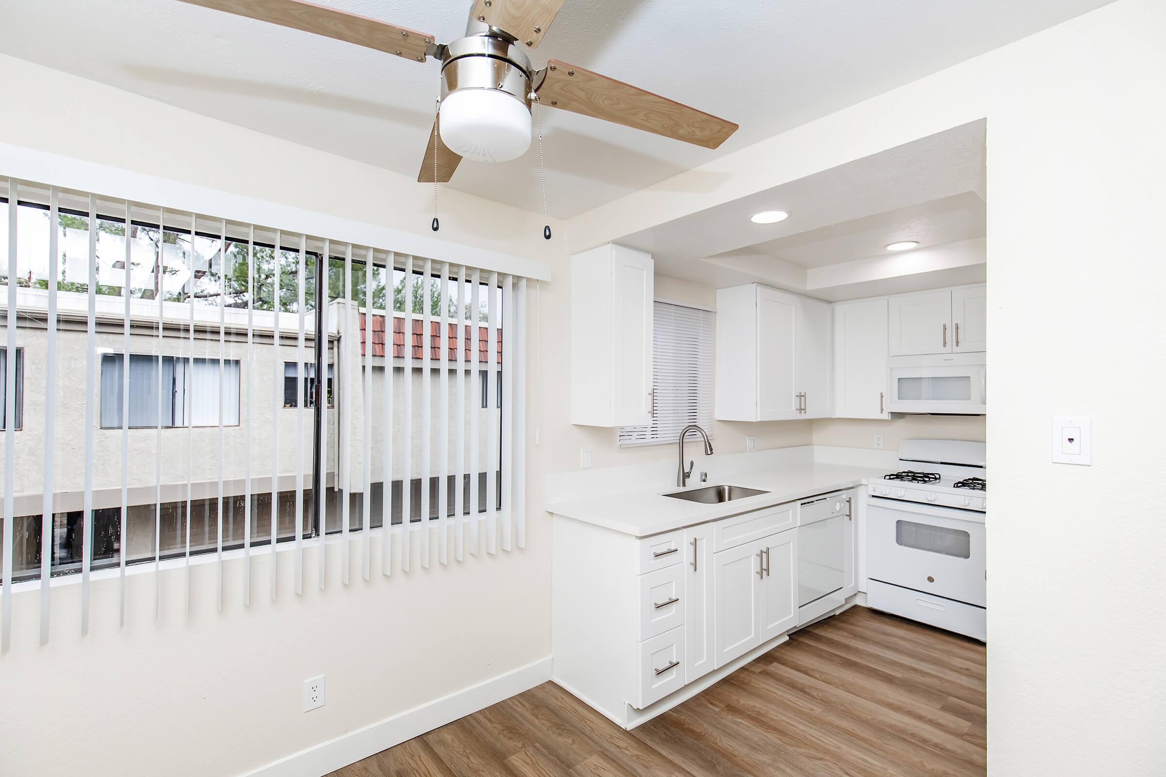 A modern kitchen featuring white cabinetry, a stainless steel sink, and a gas stove. The room has a ceiling fan and large window with vertical blinds, allowing natural light to enter. Light-colored walls and wooden flooring create a bright and inviting atmosphere.