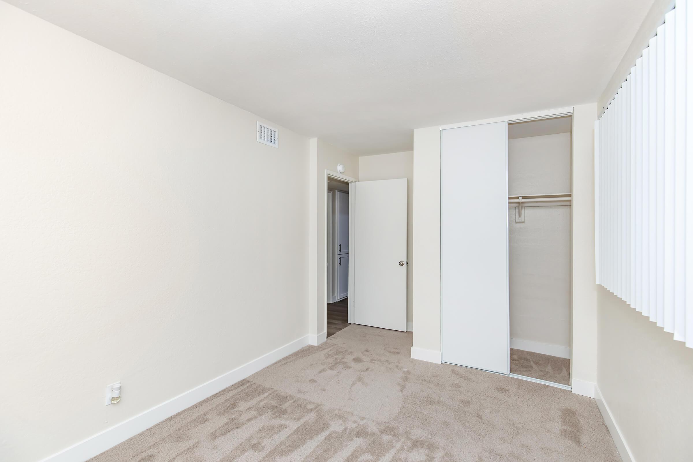 Empty, well-lit room with light-colored walls and carpet. Features a closet with sliding doors, a door leading to another room, and window coverings that allow natural light. The space appears clean and uncluttered, suitable for various uses such as a bedroom or office.