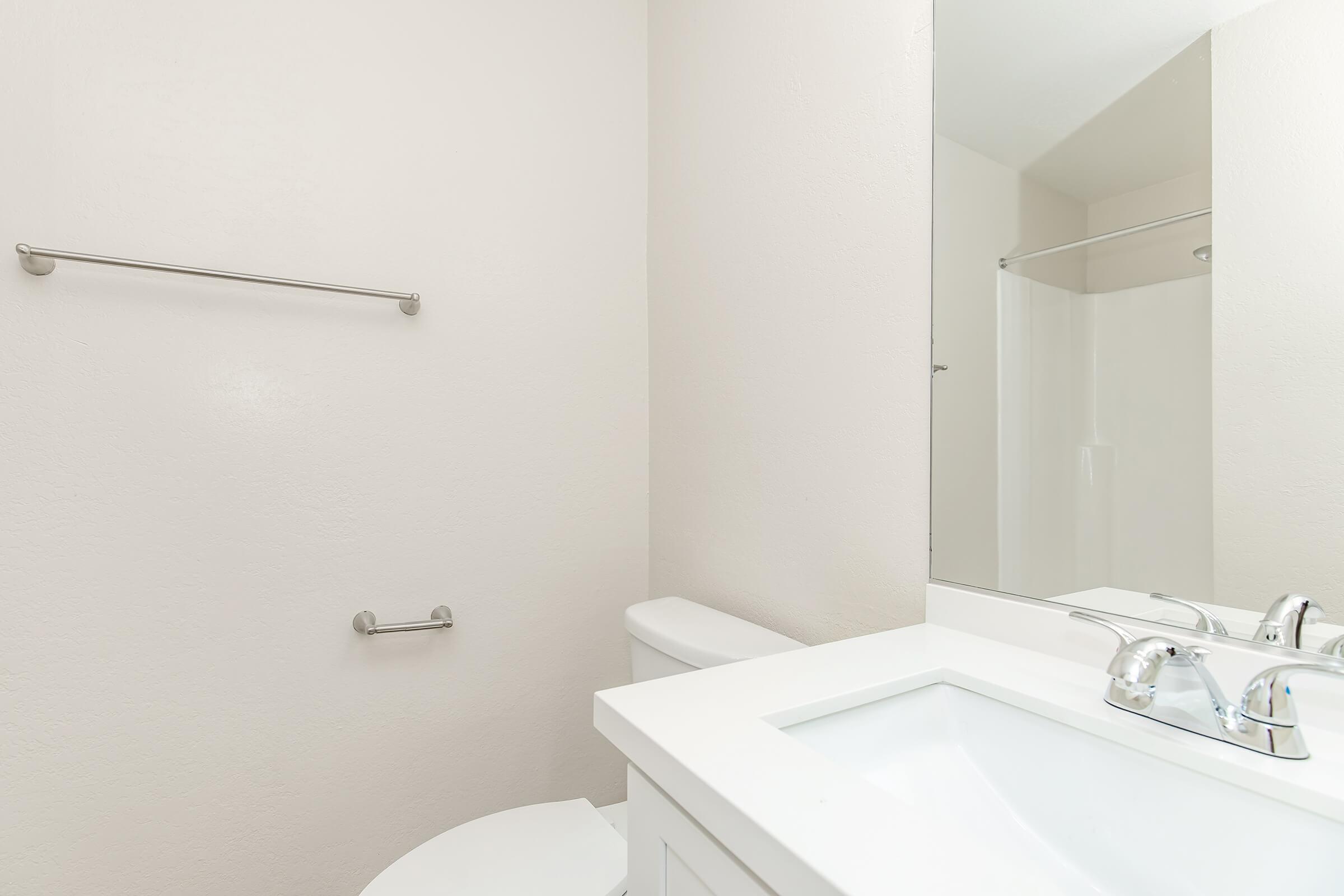 A clean and minimalistic bathroom featuring a white sink with dual faucets, a white toilet, and a walk-in shower. The walls are painted in a light color, giving the space an airy feel. A mirror above the sink reflects the simple design, with a towel bar and toilet paper holder visible.