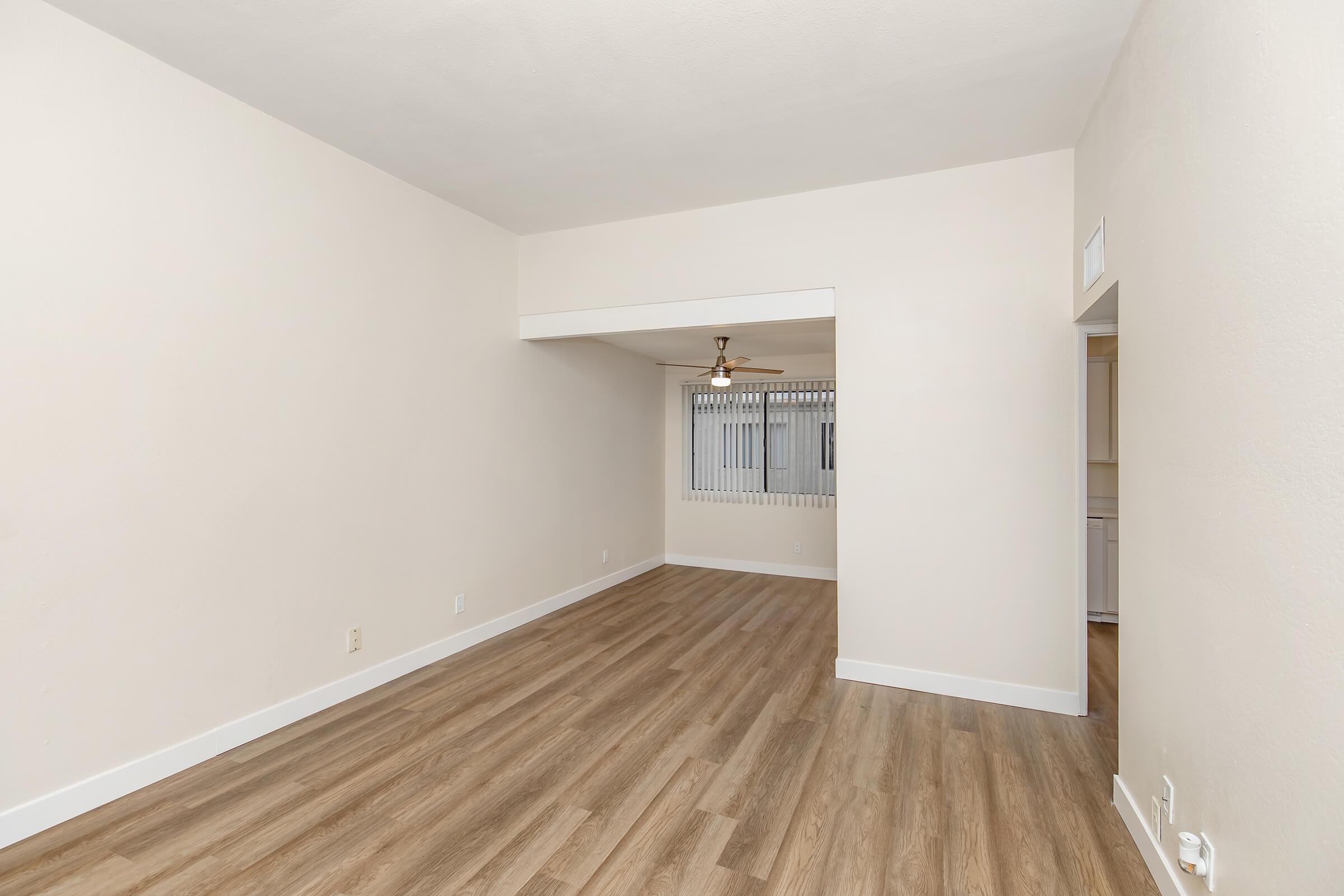 Empty room with light-colored walls and hardwood flooring. A ceiling fan is visible, and there's a small archway leading to another space with a window covered by vertical blinds. The overall ambiance is bright and airy, suitable for decoration or furnishing.