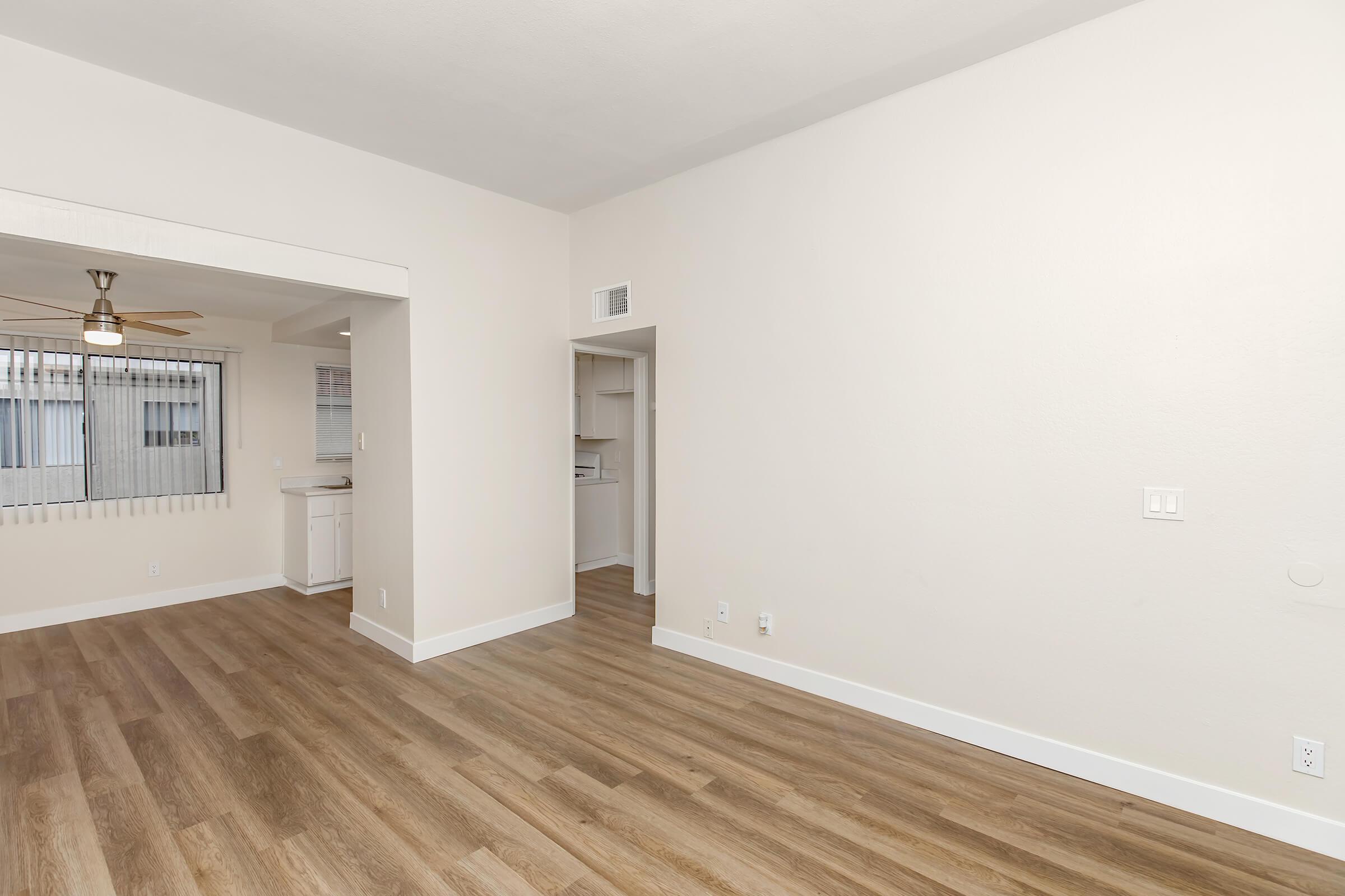 A spacious, light-filled room with light-colored walls and wooden flooring. On the left, there is an open doorway leading to a kitchen area with a ceiling fan. The room is uncluttered, with large windows allowing natural light, creating a welcoming and airy atmosphere.