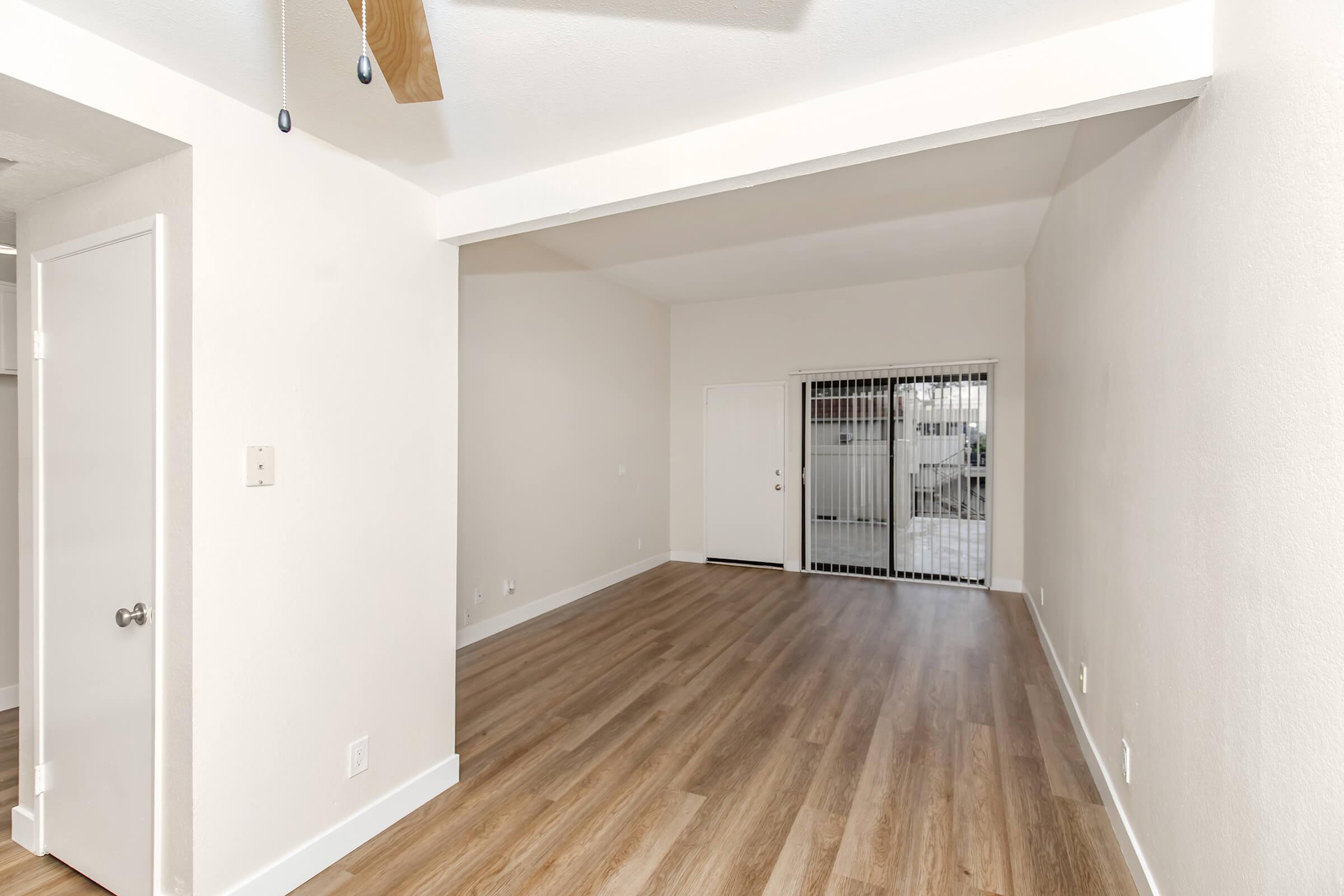 A bright and spacious interior with light-colored walls and smooth wooden flooring. There is a doorway leading outside, and a ceiling fan mounted above. The area is well-lit and uncluttered, creating an inviting atmosphere.