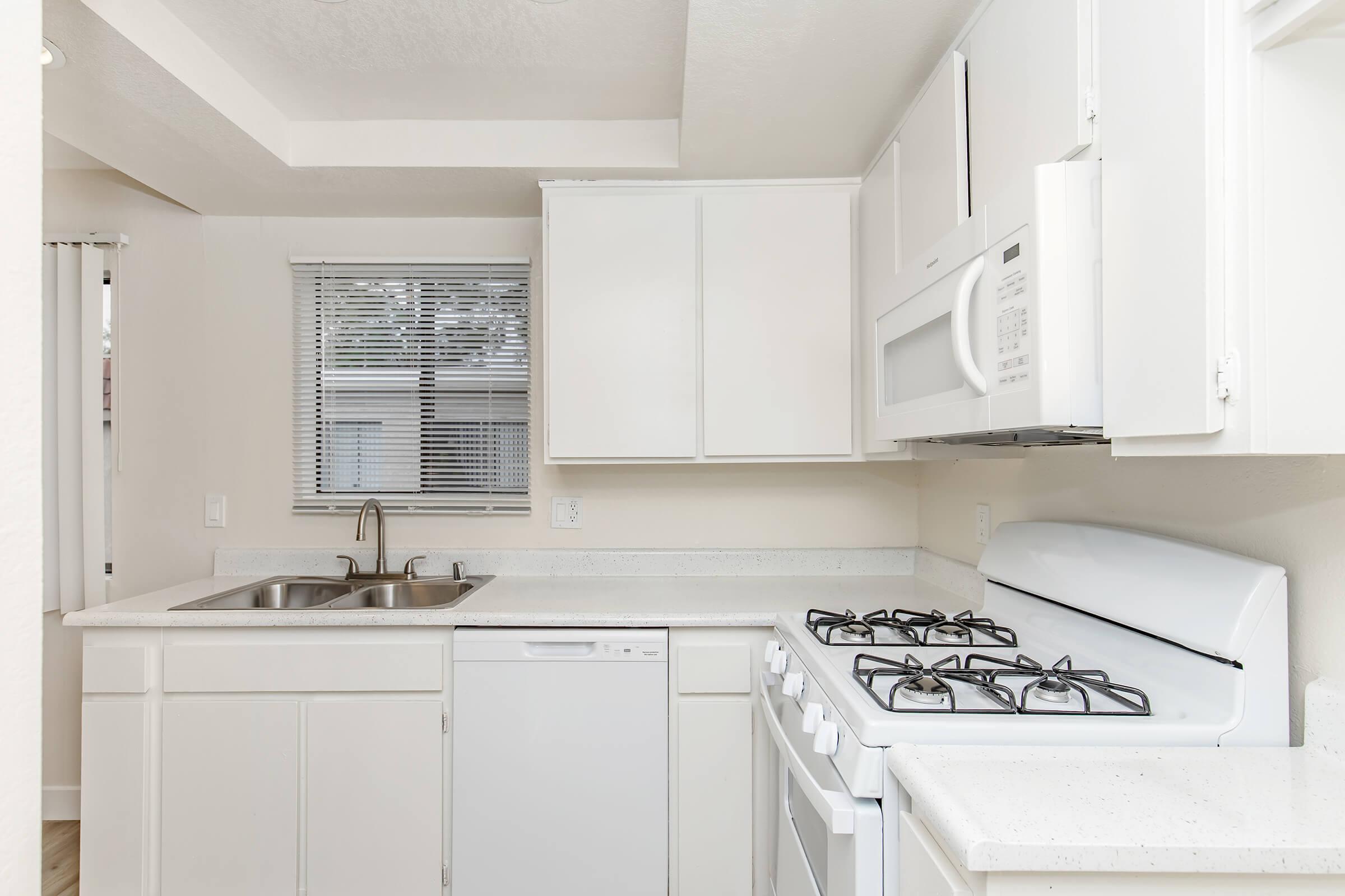 Modern kitchen featuring white cabinetry, a stainless steel sink, a dishwasher, and a gas stove. The countertop is light-colored, and there is a microwave mounted above the stove. A window with blinds allows natural light in, creating a bright and clean atmosphere.