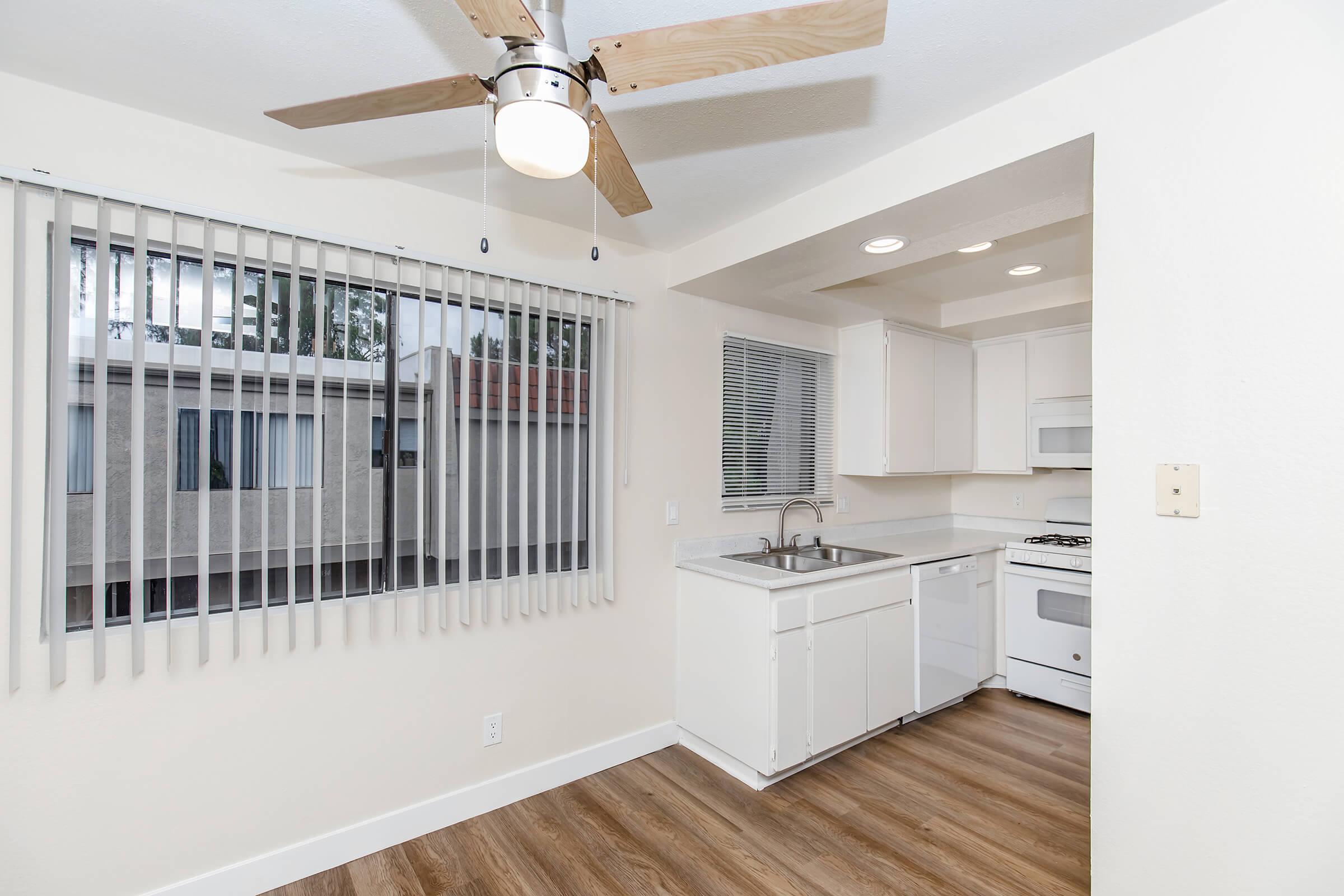 A bright, modern kitchen featuring a white countertop, sink, and appliances. A ceiling fan with wooden blades is installed, and large windows with vertical blinds allow natural light to enter. The flooring is a warm wood texture, creating a welcoming atmosphere.